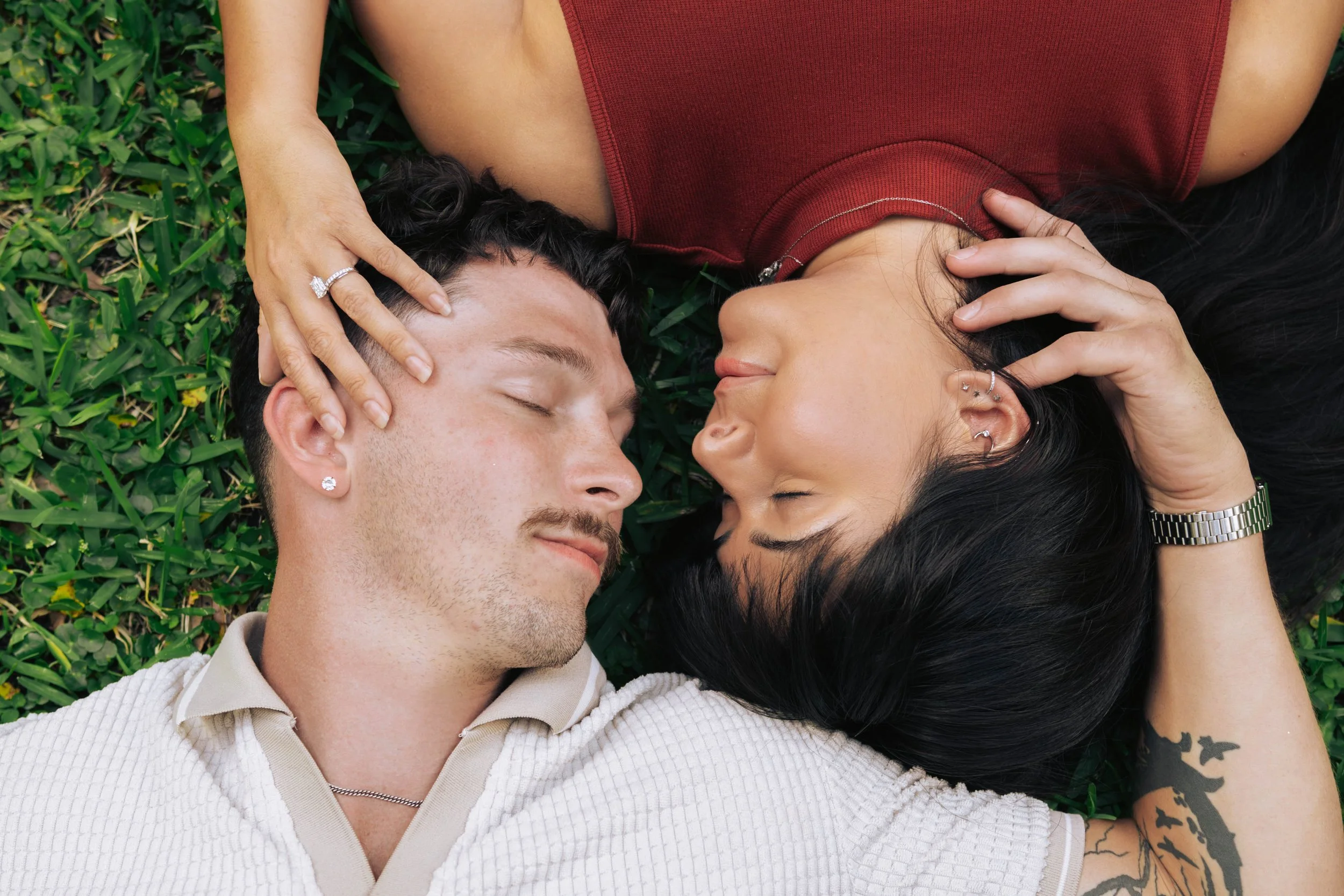 A man and a woman lying on the grass, facing each other with their eyes closed, touching each other's hair gently.