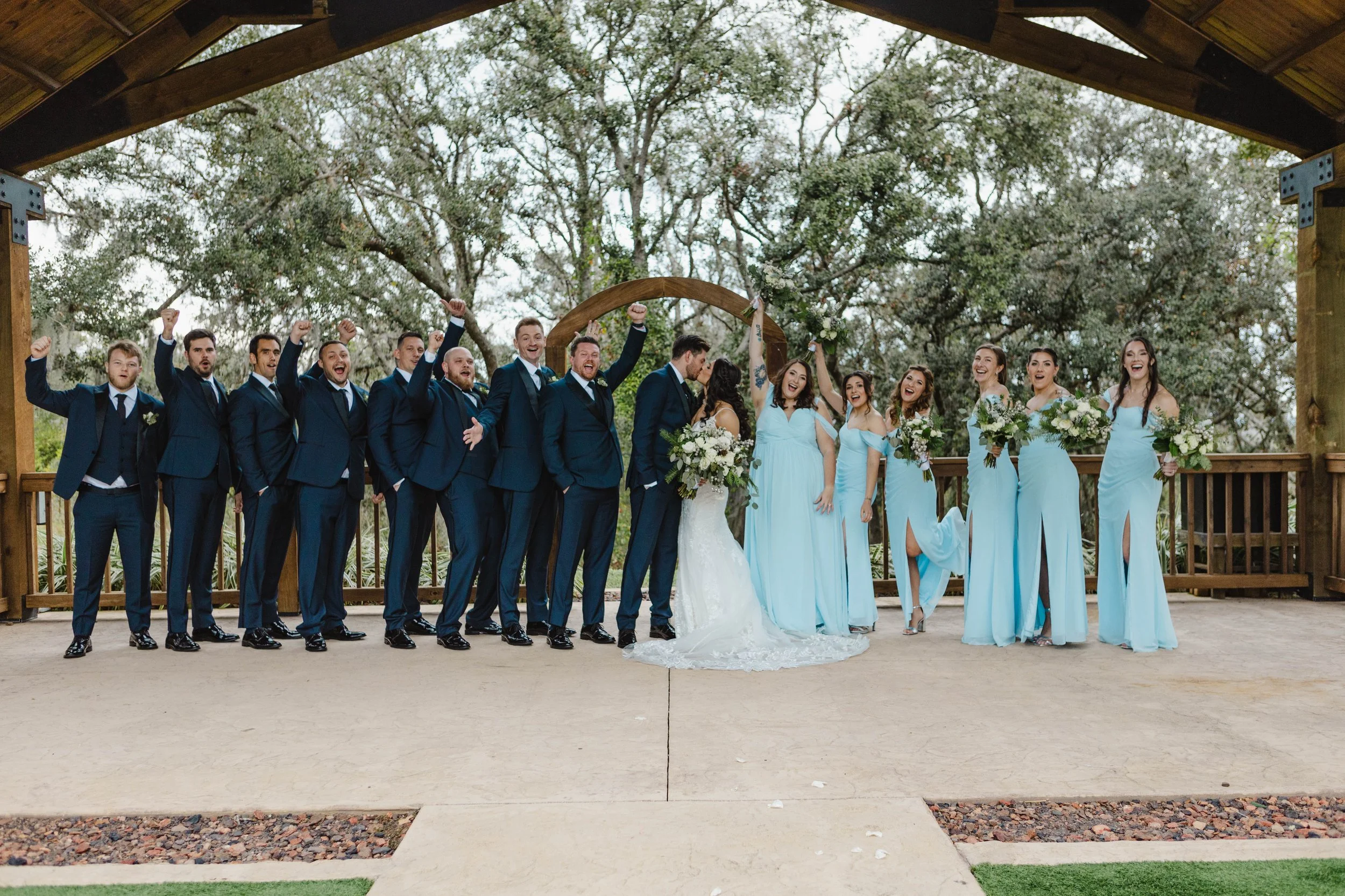 A group of people dressed in wedding attire standing on a wooden deck outdoors, celebrating under a wooden structure with trees in the background. The group includes groomsmen in navy suits and bridesmaids in light blue dresses, with the bride and gr
