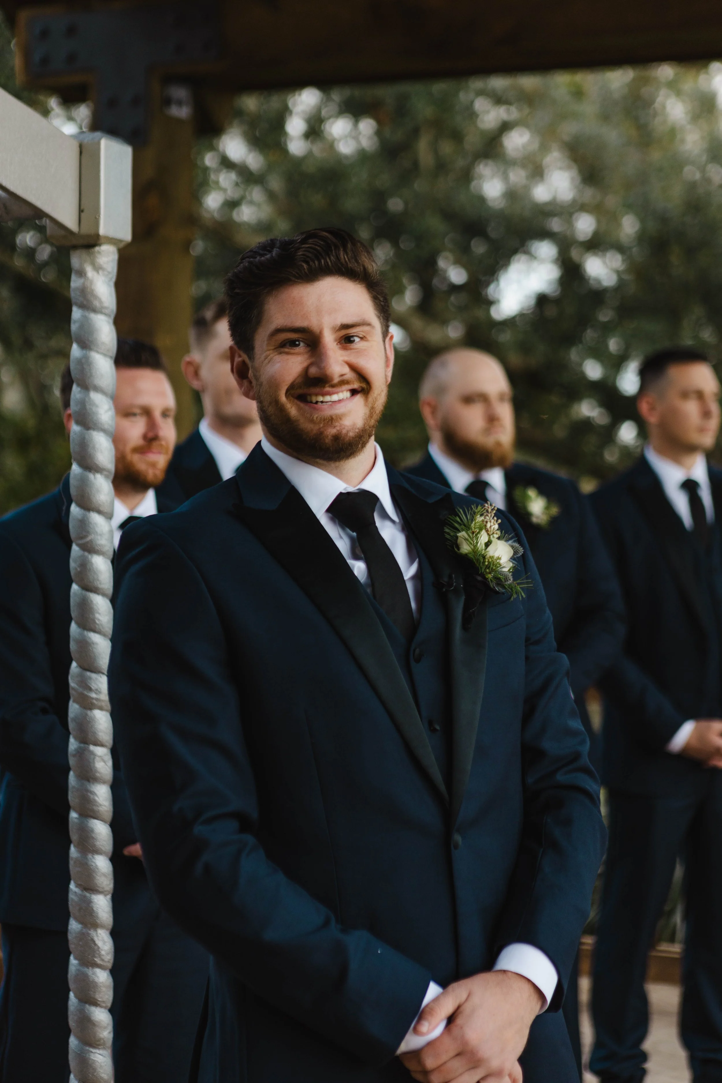 A group of men in formal suits standing outdoors, likely during a wedding or formal event. The man in the front is smiling, with dark hair and a beard, wearing a navy suit with a white shirt and a boutonniere. Other men are visible in the background,