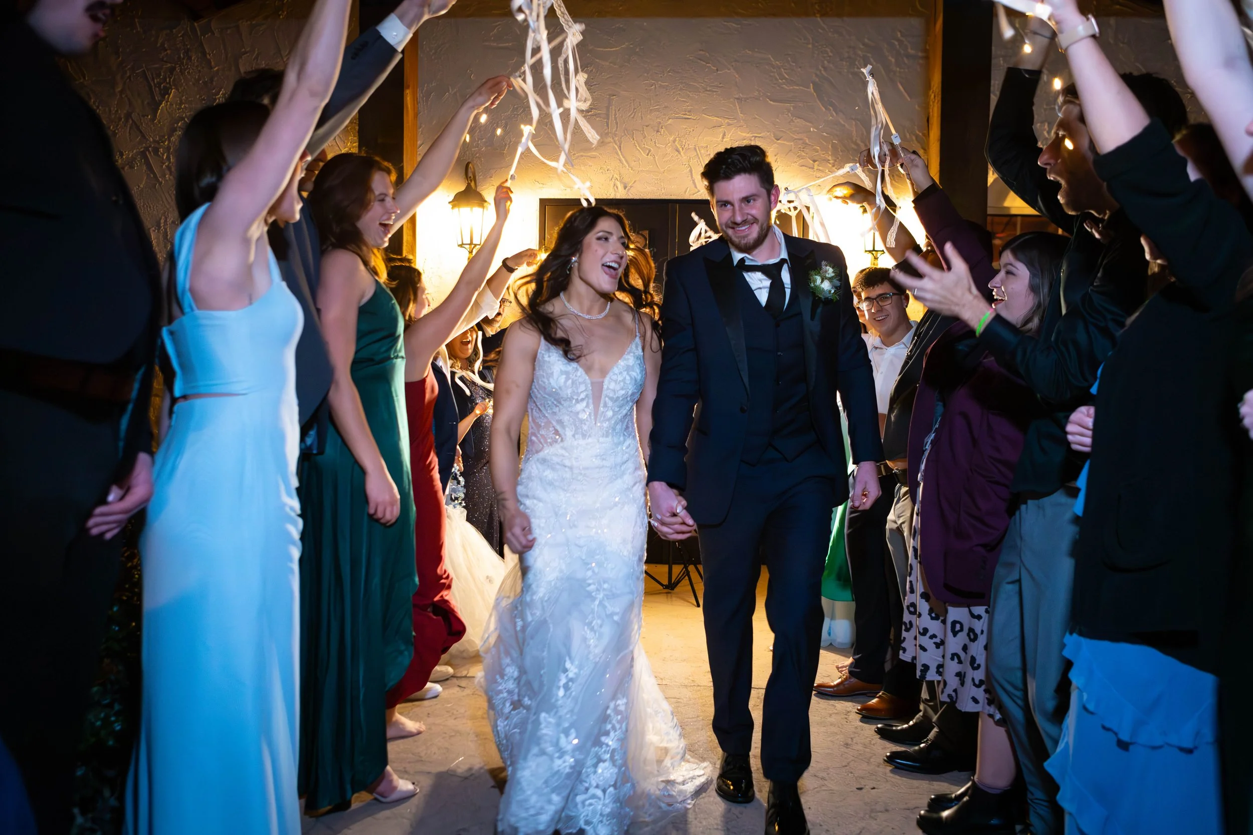 A bride and groom holding hands in the center, surrounded by friends and family celebrating at their wedding reception. Everyone is smiling and raising their hands, with streamers hanging down from the ceiling.