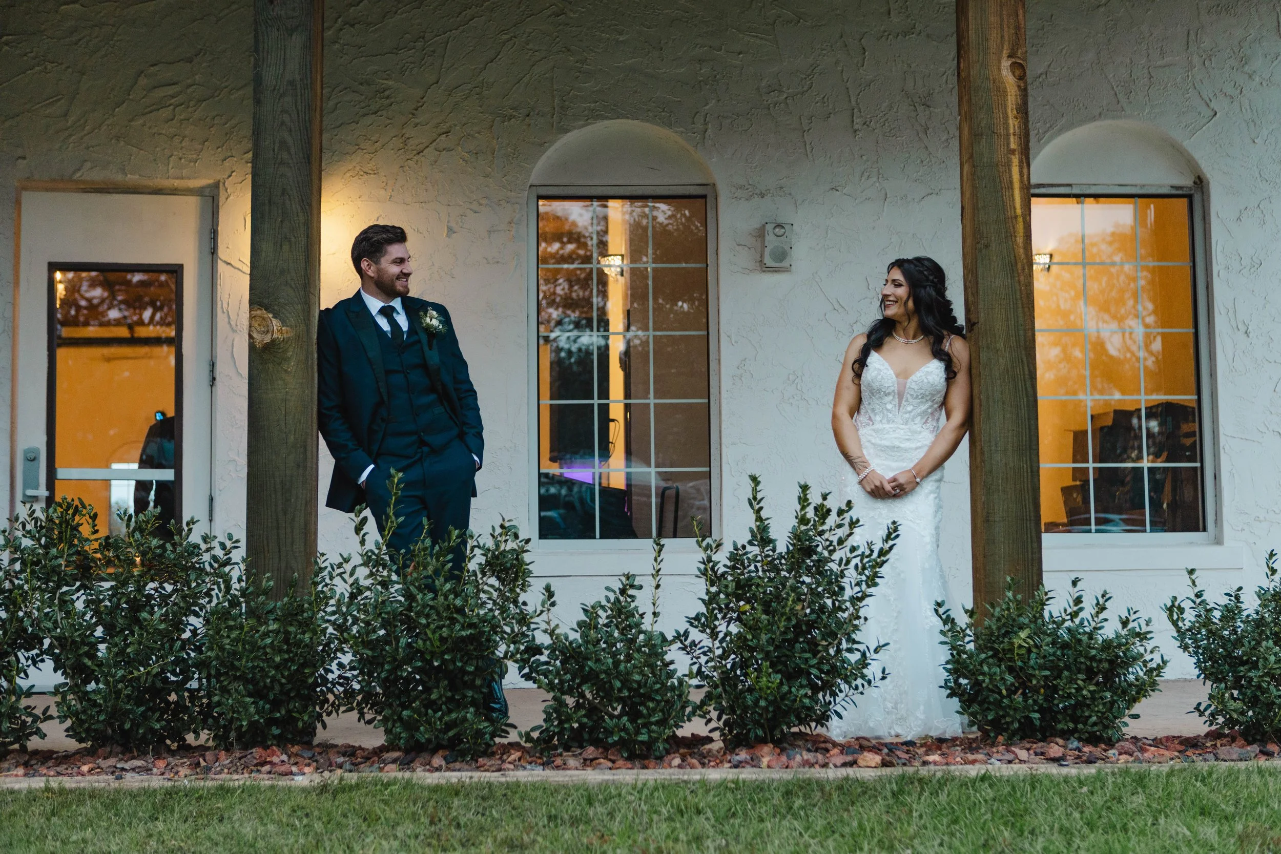 A bride in a white wedding dress and a groom in a dark suit are standing outside a building with textured white walls and large windows, smiling at each other during sunset.