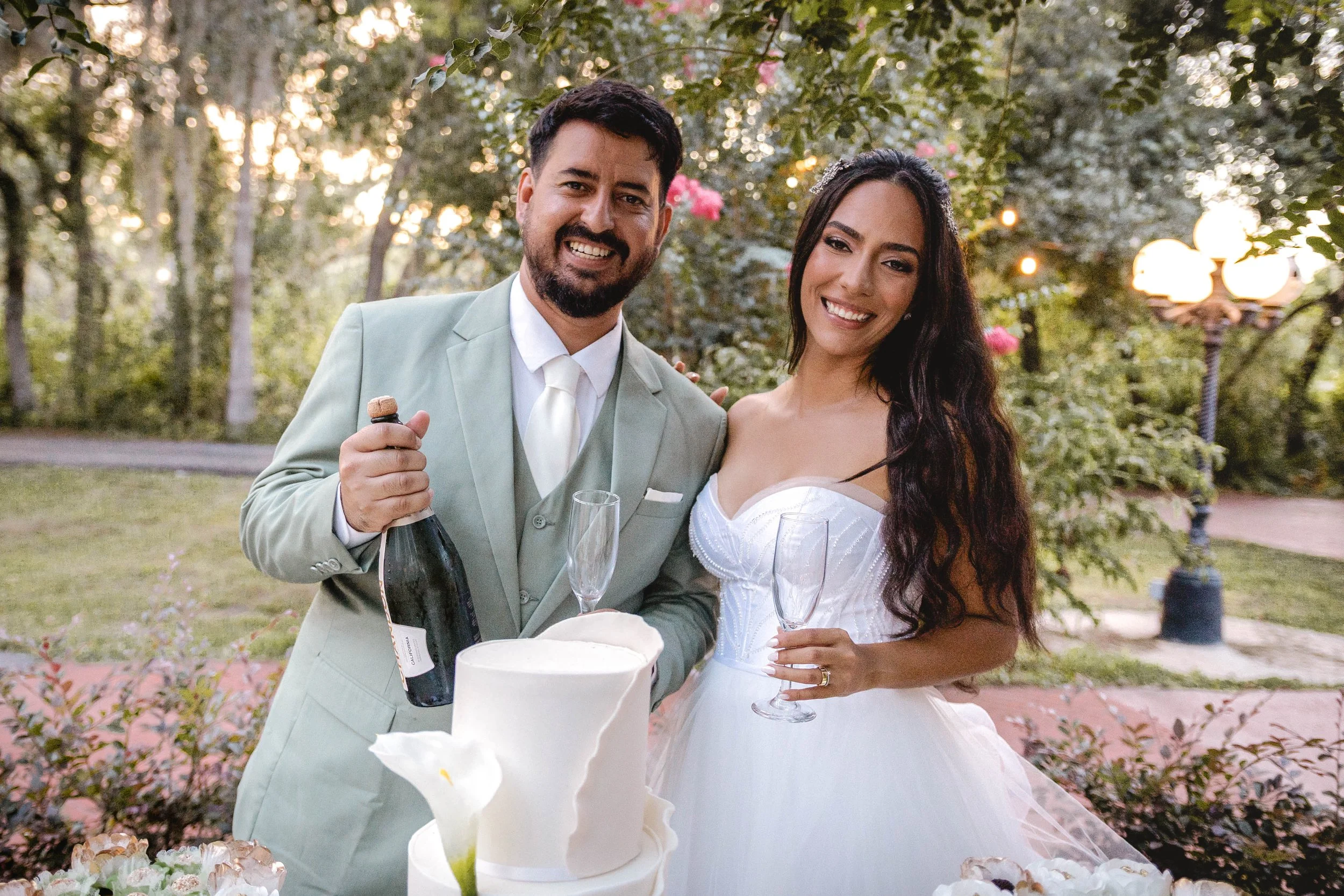 A newlywed couple smiling and celebrating outdoors, holding champagne glasses near a wedding cake, with trees and warm lighting in the background.