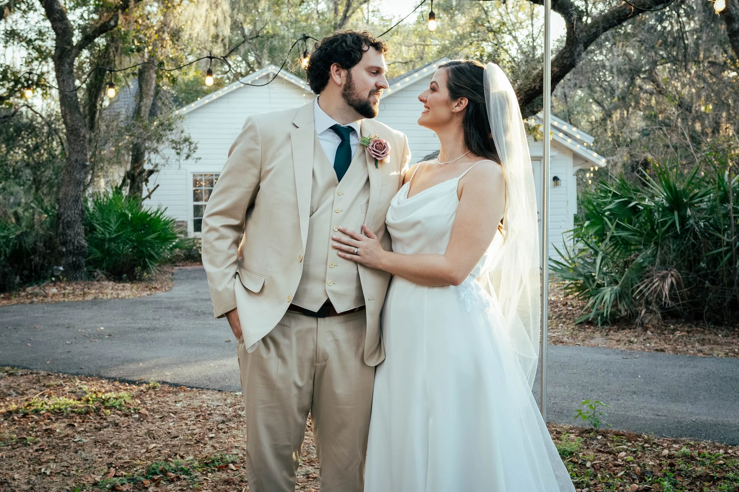A bride and groom standing close, smiling and looking into each other's eyes during their outdoor wedding at sunset, with string lights and trees in the background.