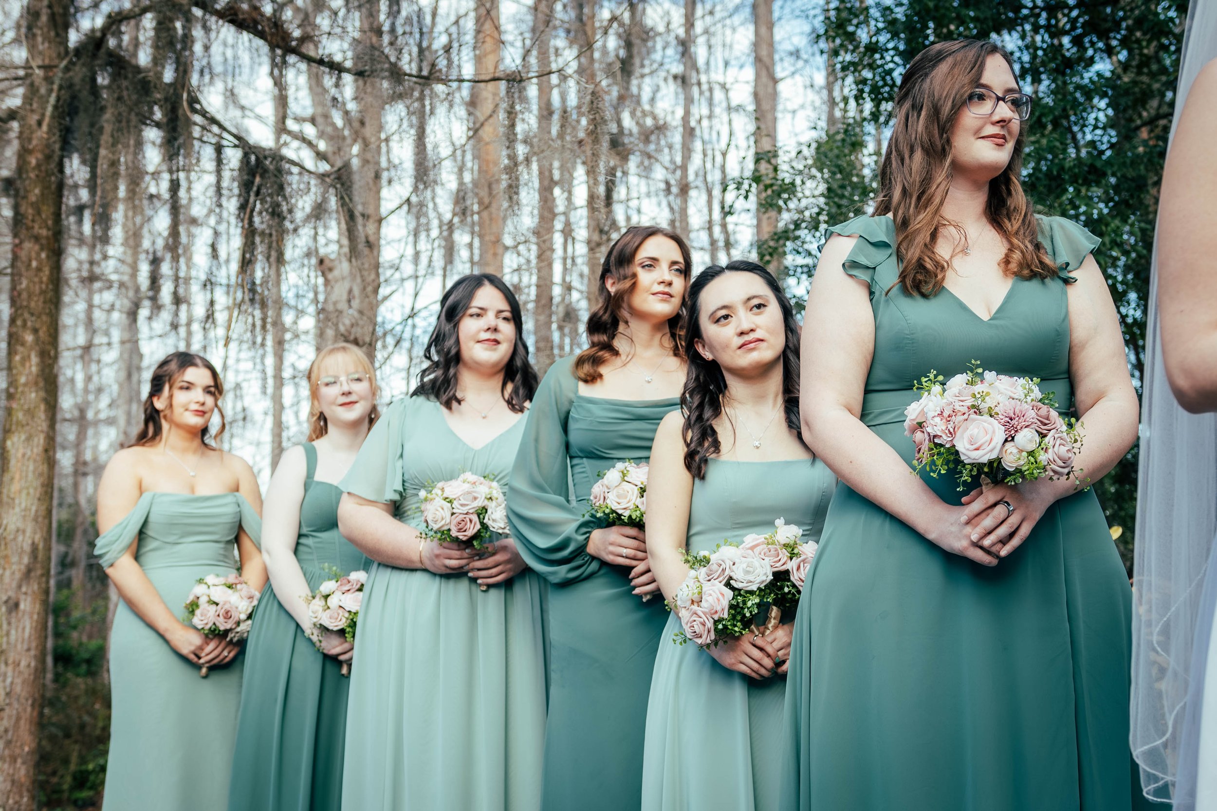 A group of women in matching sage green dresses are standing in a line outdoors, each holding a bouquet of light pink and white roses. They appear to be part of a wedding or formal event, set against a background of tall trees and a bright, partly cl