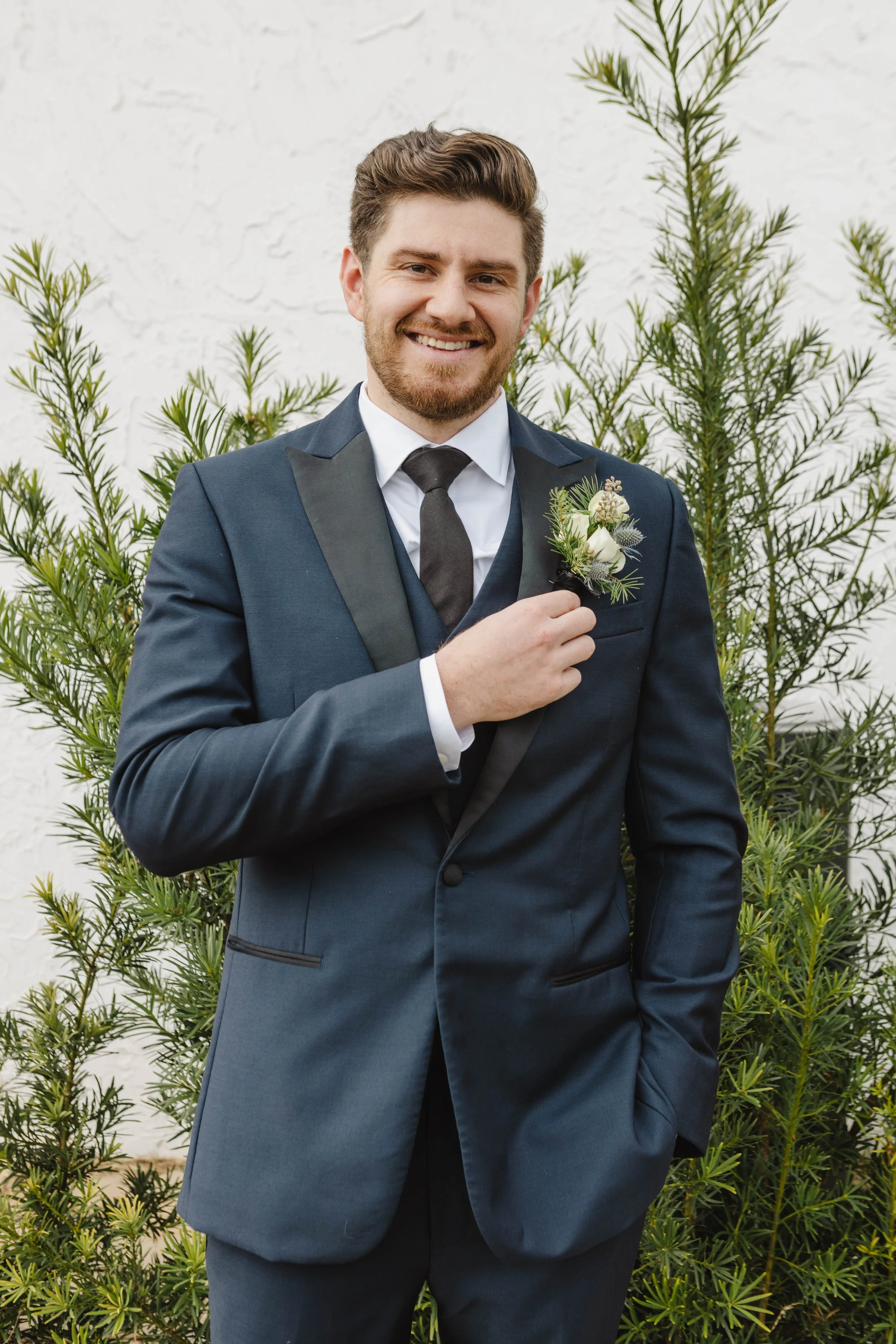 A young man in a formal navy blue suit, white shirt, and black tie, smiling and holding a boutonniere on his suit's lapel, standing in front of a white textured wall and green shrubbery.