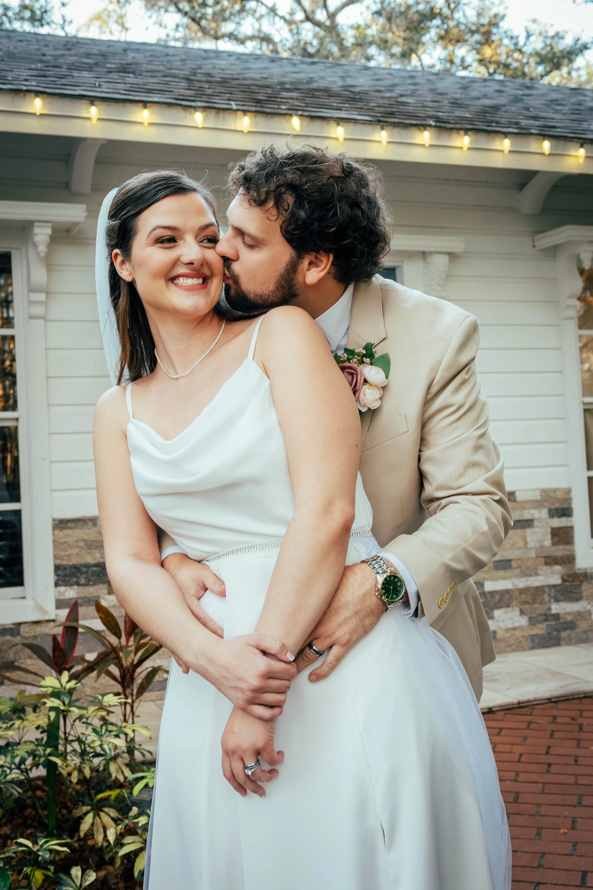 A newlywed couple, a woman in a white wedding dress and a man in a beige suit, sharing a kiss and smiling in front of a house with string lights and plants.