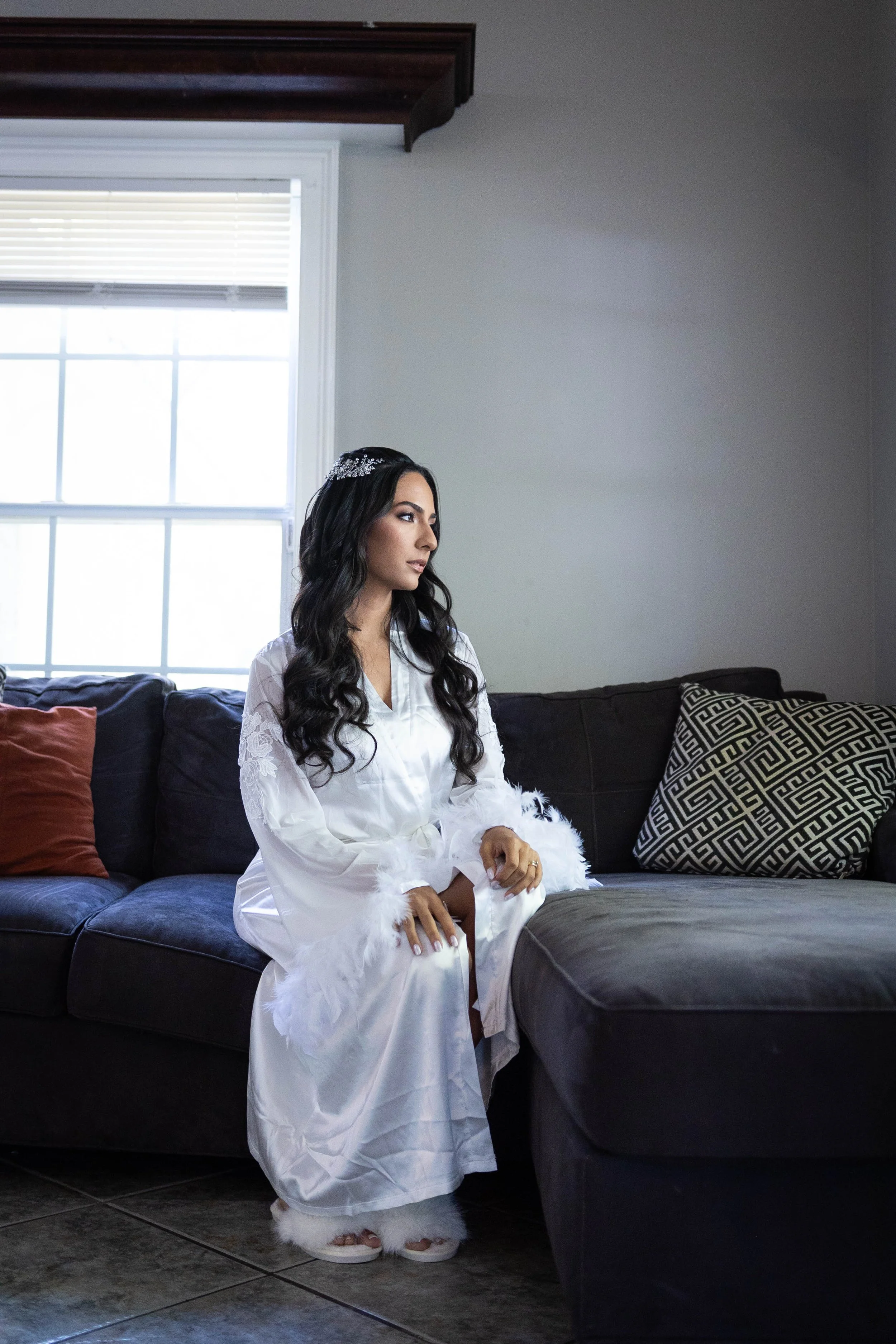 A woman with long, dark hair sitting on a dark gray couch in a living room, wearing a white satin robe with feather trim and a tiara, and looking to the right. The room has gray walls, a window with blinds, and decorative pillows on the couch, includ