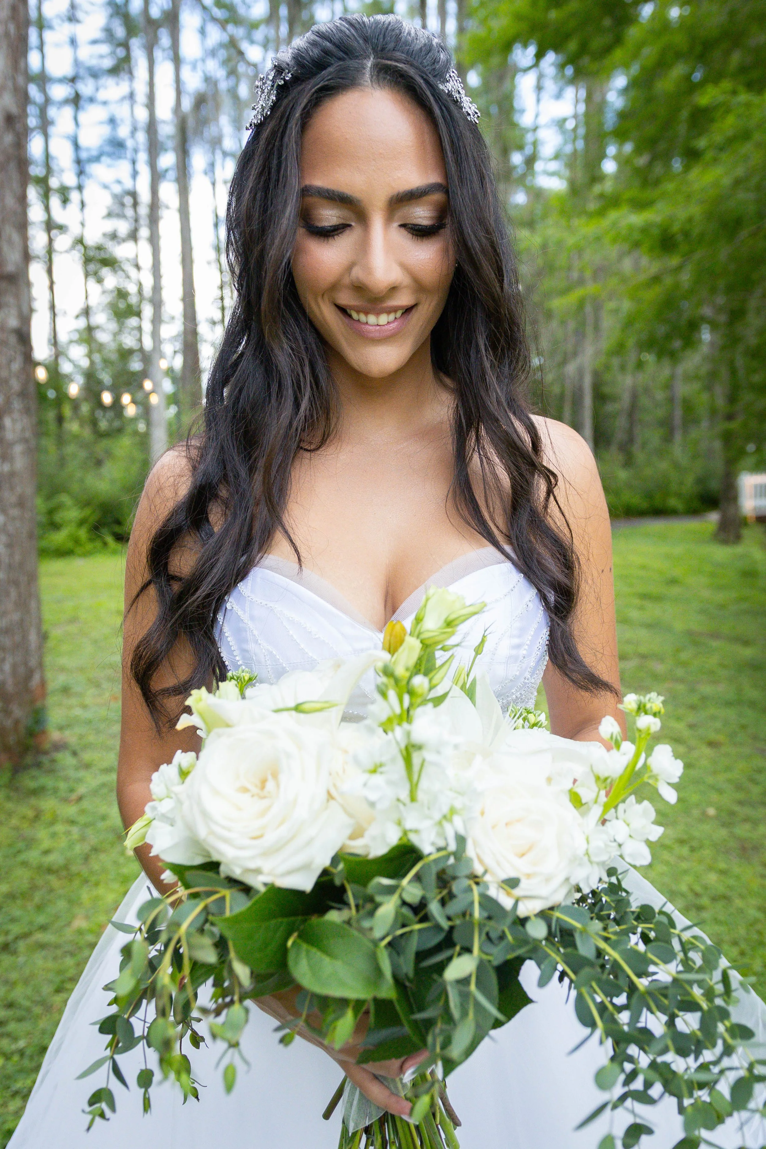 A woman in a white wedding gown holding a large bouquet of white flowers and greenery outdoors in a wooded area.