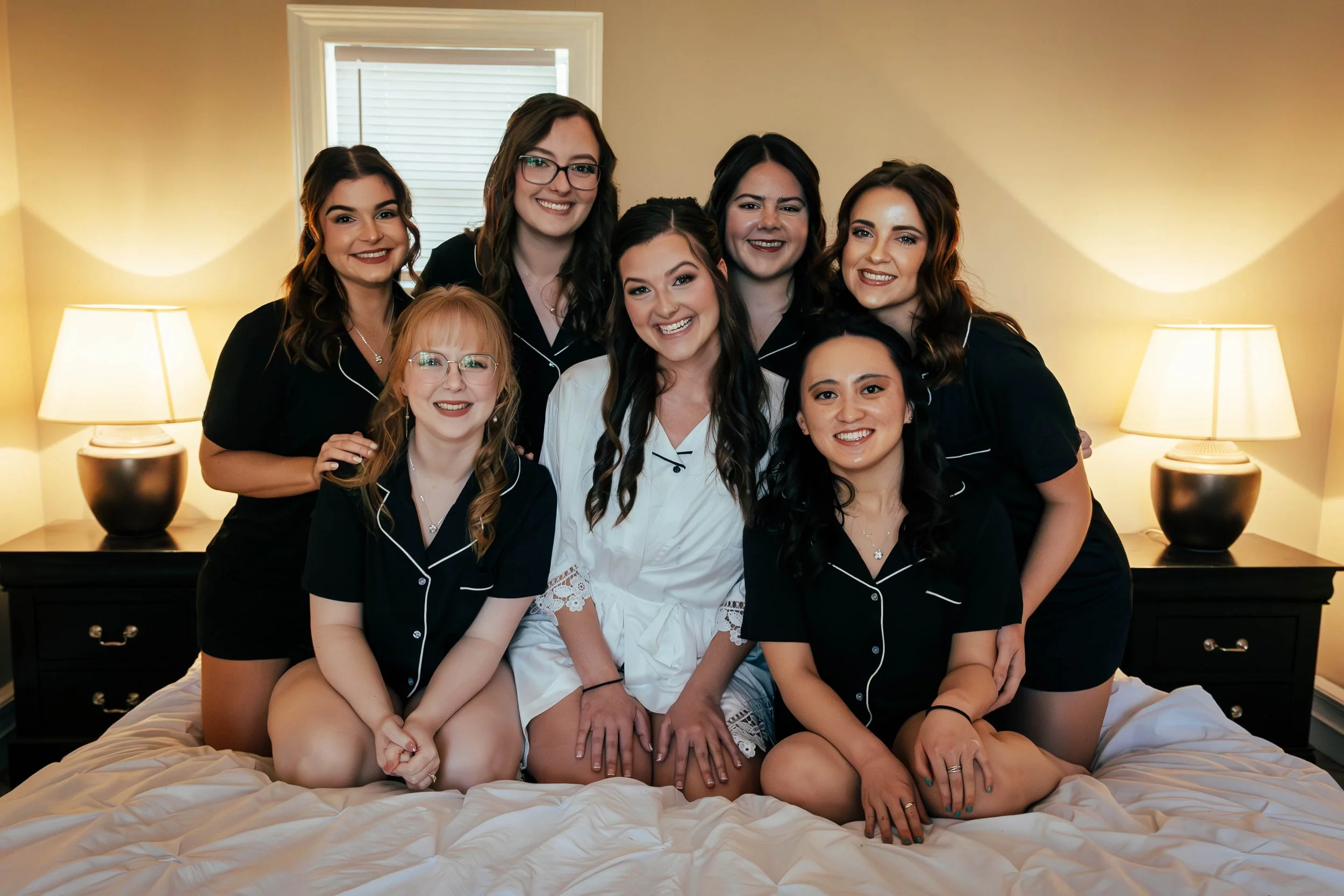 Group of women in pajamas sitting and kneeling on a bed in a bedroom, smiling at the camera with lamps on bedside tables and a window with blinds behind them.