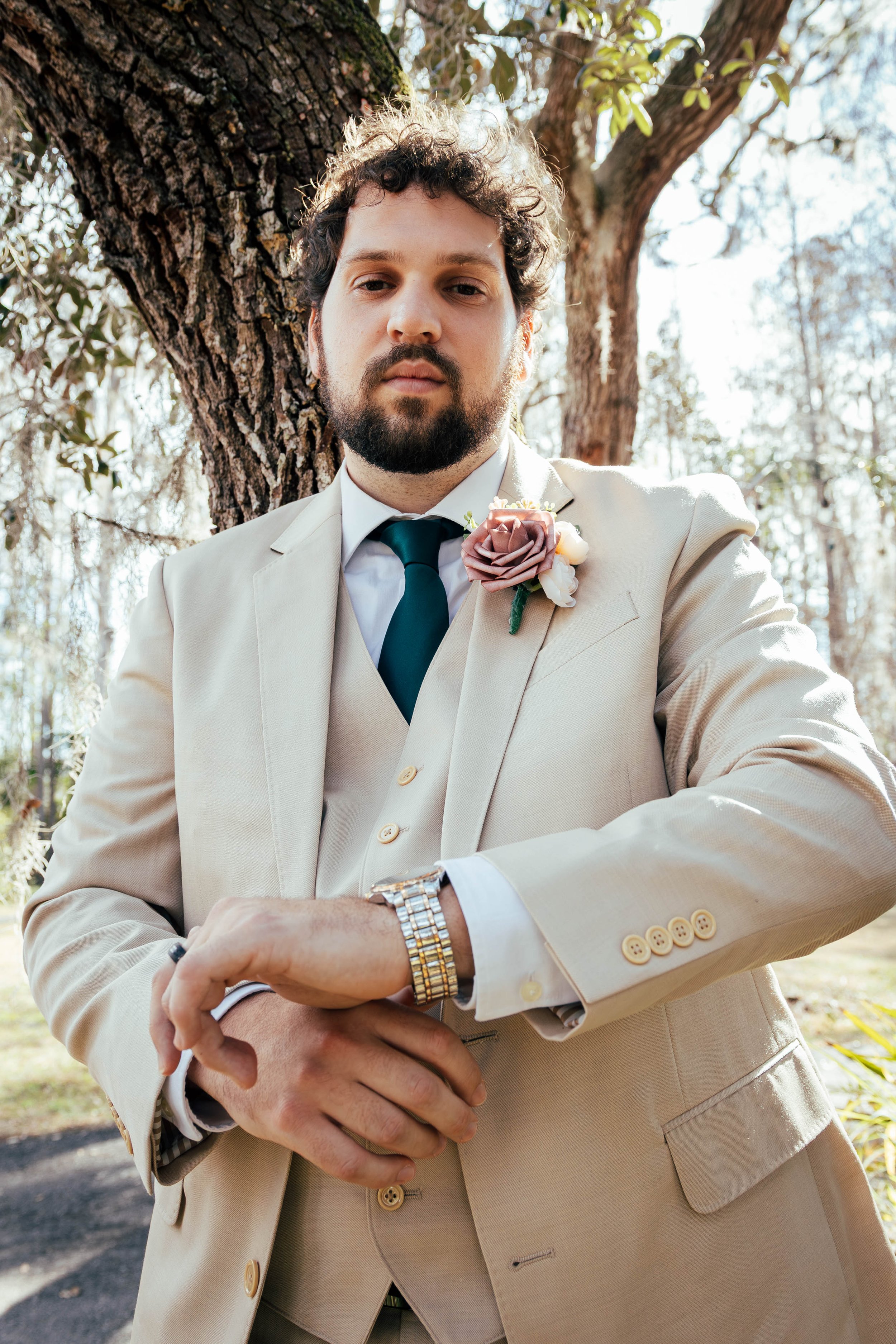 A man with curly dark hair, a beard, and mustache wearing a beige suit and teal tie, standing outdoors near a tree. He has a floral boutonniere on his lapel and is wearing a gold watch and ring, with a serious expression.