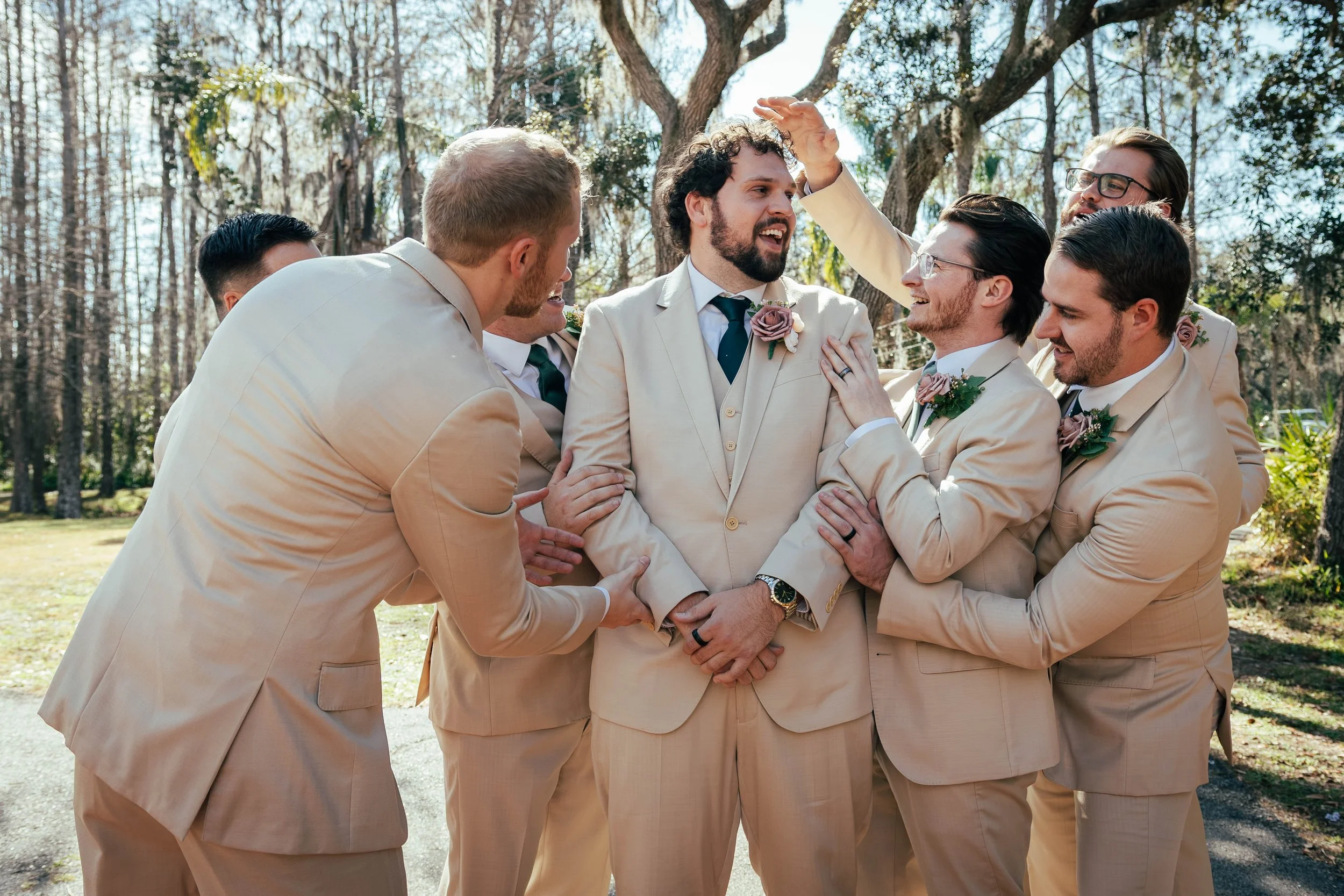 Group of men dressed in beige suits, standing outdoors, smiling and embracing, with trees and blue sky in the background.