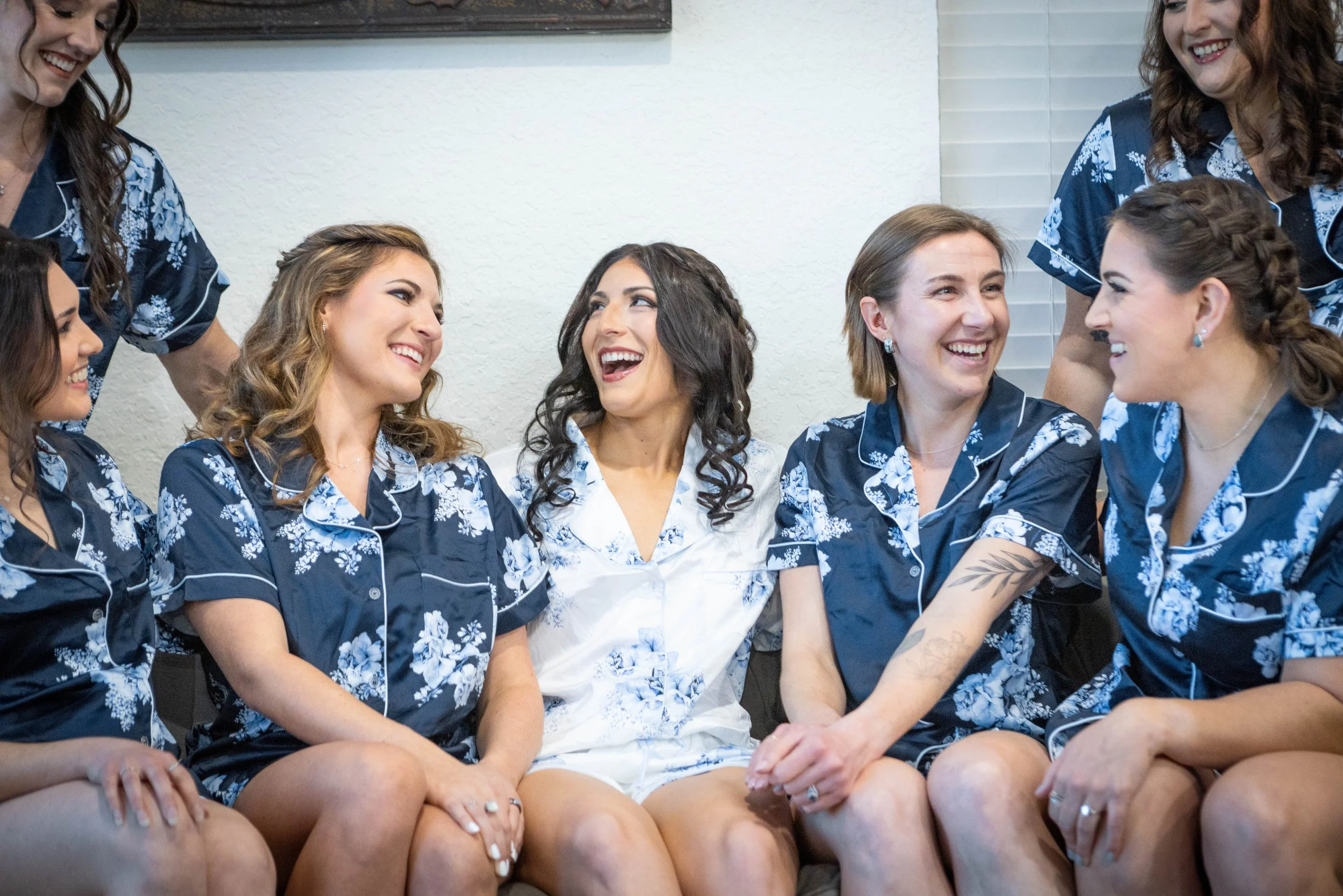 Six women sitting closely together, wearing matching blue floral pajamas, smiling and looking at each other in a cozy indoor setting.