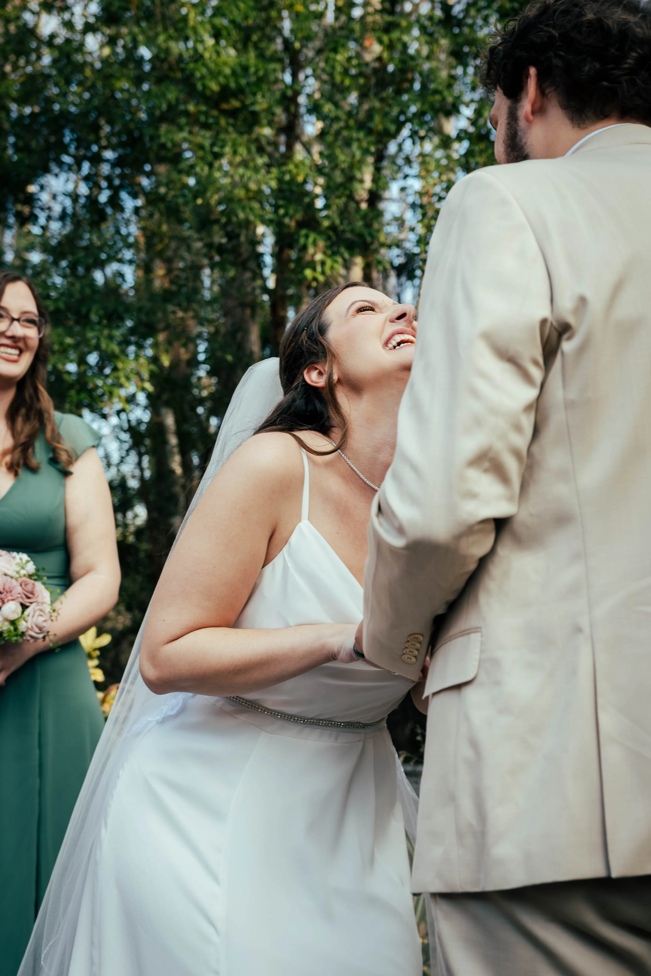 A woman in a white wedding dress is smiling and looking up at a man in a beige suit during a wedding ceremony outdoors, with another woman in a green dress holding a bouquet of flowers in the background.