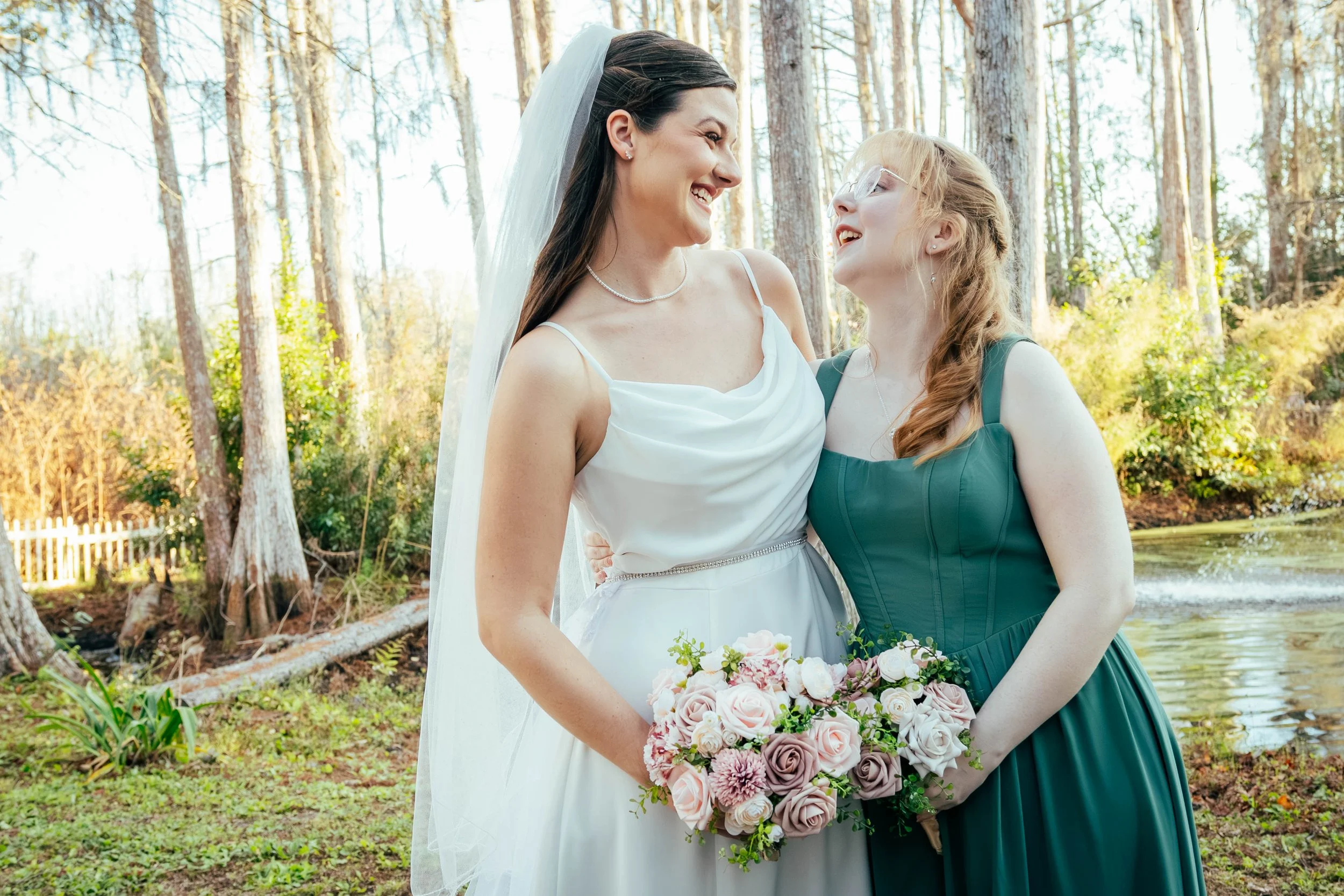 Two women, one in a white wedding dress and veil, and the other in a dark green dress, smiling and looking at each other outdoors in a wooded area, holding a bouquet of pink and white flowers.