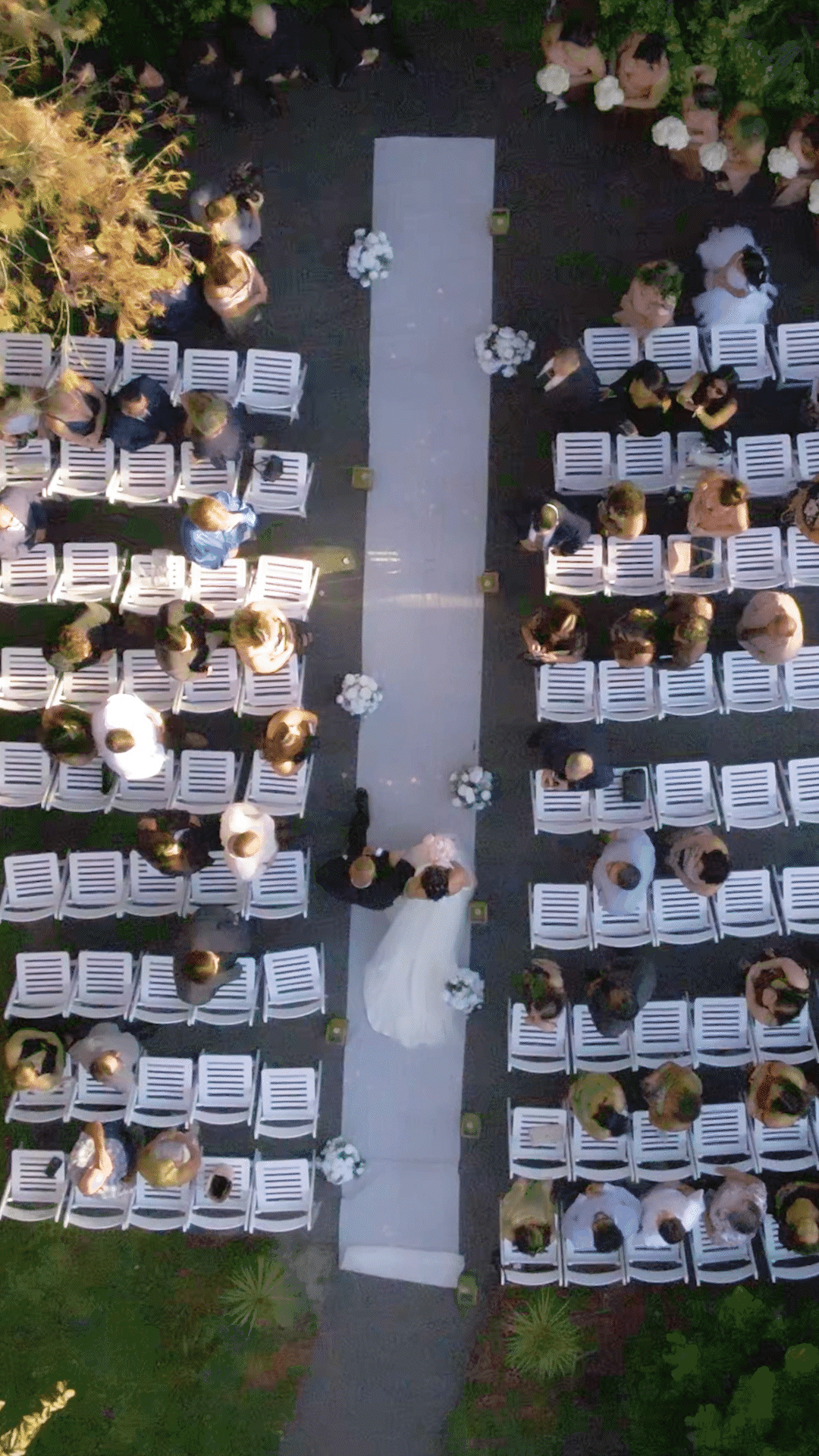 Aerial view of a wedding ceremony with rows of white chairs arranged on both sides of a gray aisle, decorated with white flower arrangements, with guests seated and some standing.