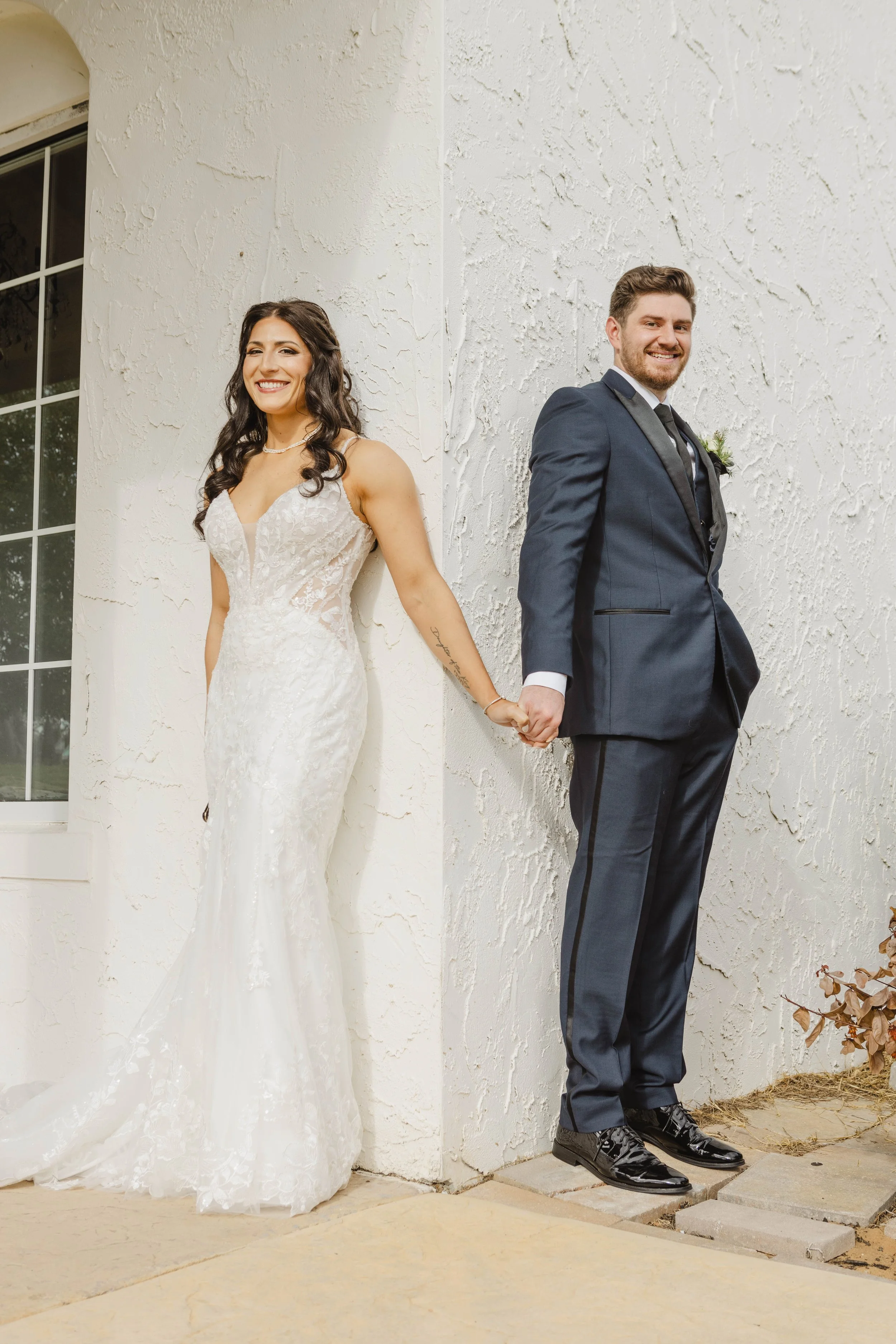 A bride and groom standing apart, holding hands and leaning against a textured white wall outside a building with a window. The bride wears a white lace wedding gown with a V-neckline, and the groom wears a dark suit with a tie and boutonniere. They 