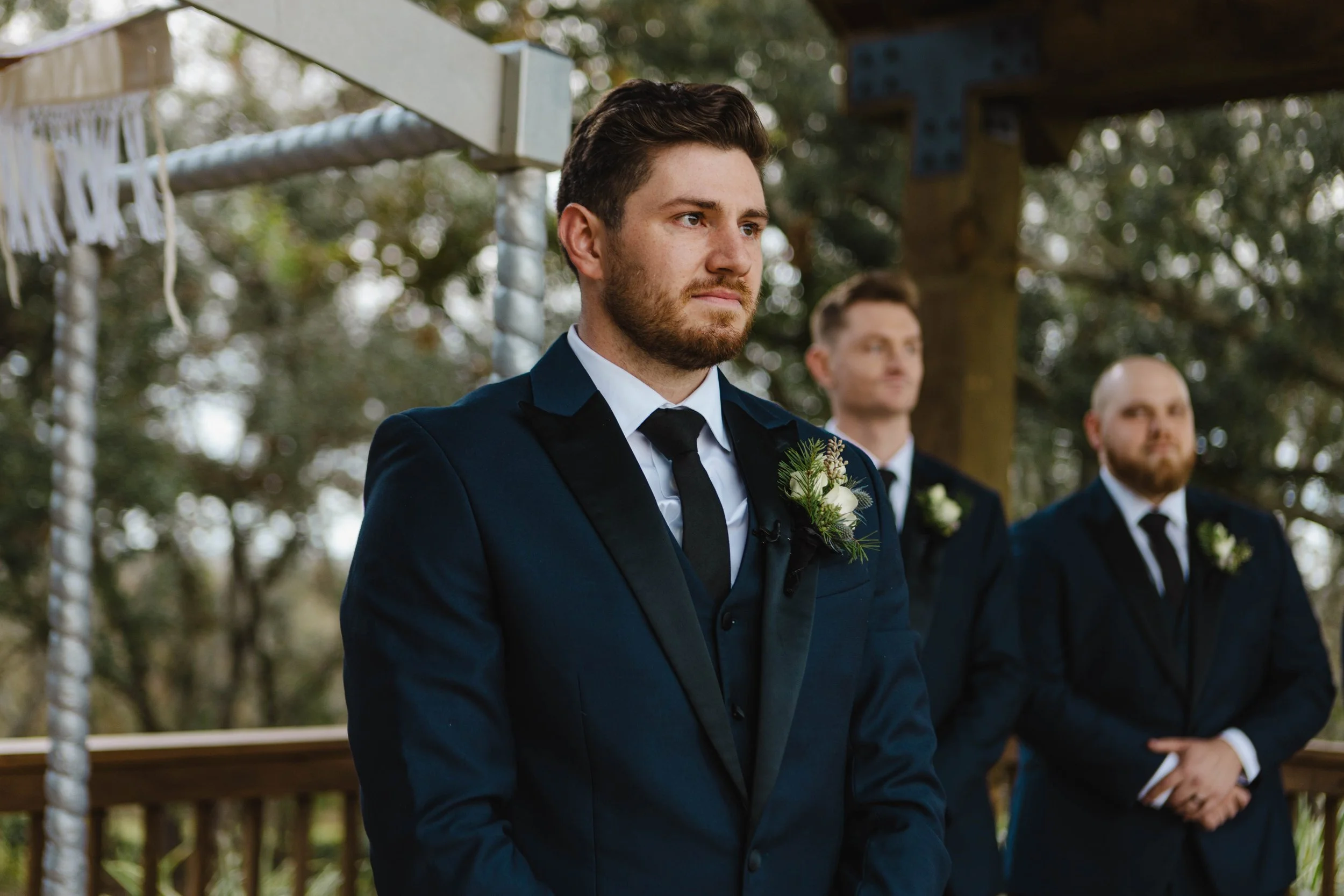 A groom with a beard and brown hair, wearing a dark blue tuxedo with a white shirt, black tie, and boutonniere, standing outdoors during a wedding ceremony.