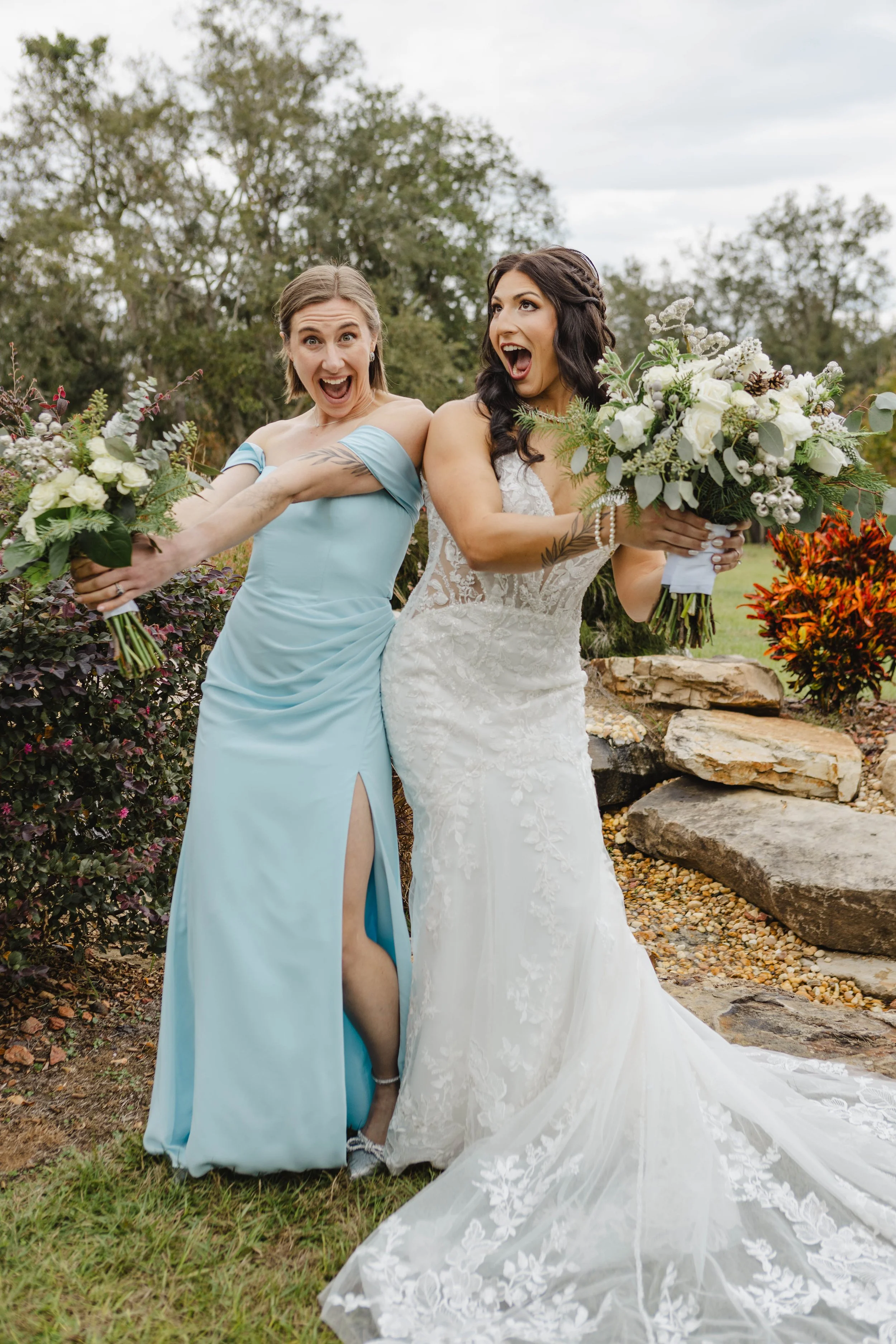 Two women, one in a light blue dress and the other in a wedding gown, are joyfully posing outdoors with big smiles. They are holding bouquets of flowers and seem excited. The background features greenery, trees, and a cloudy sky.