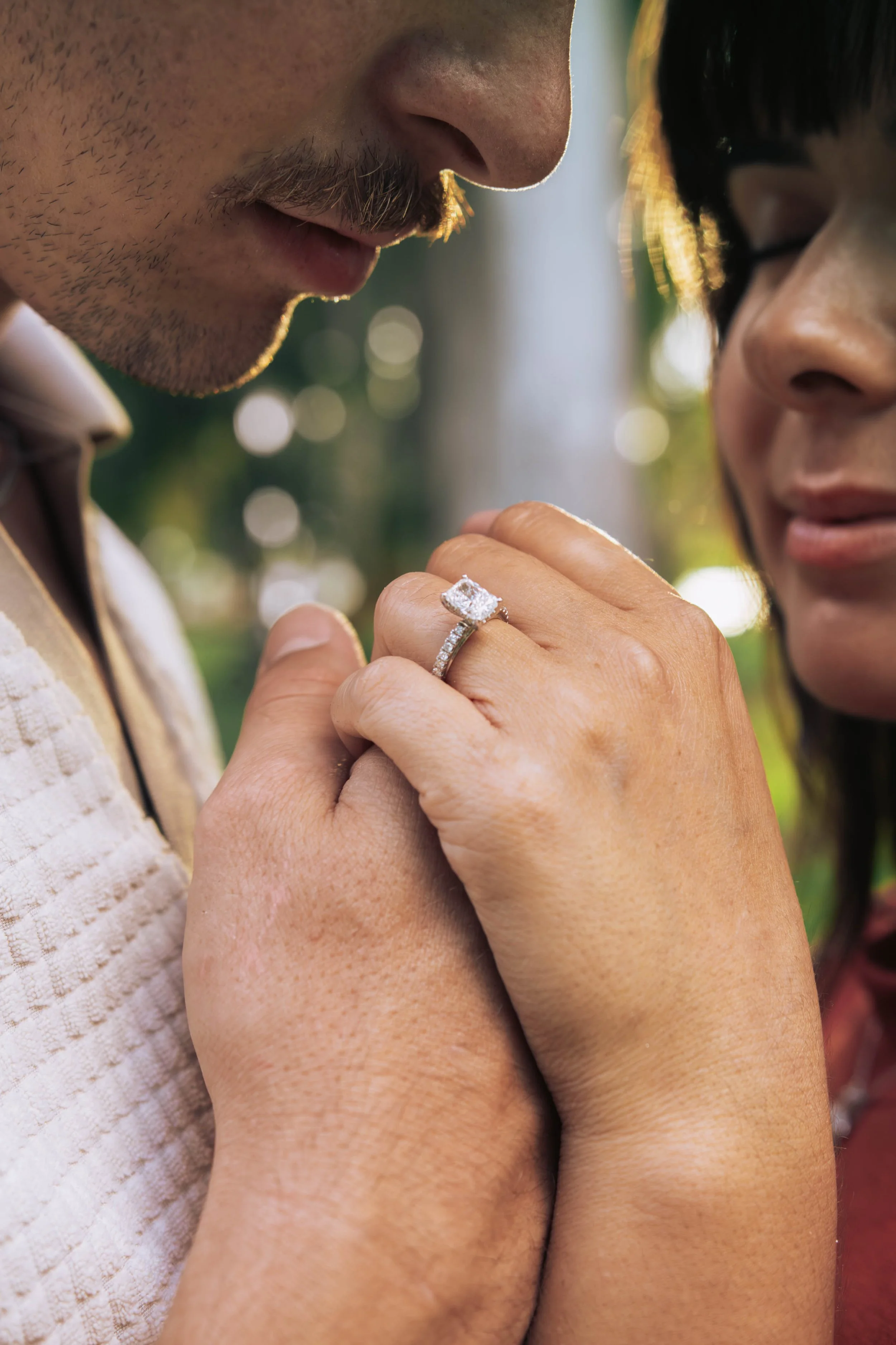 A couple is close together, with the woman holding a diamond engagement ring up to the man's face. The man has a beard and mustache, and the woman is wearing glasses and has dark hair. They are outdoors with blurred natural background.