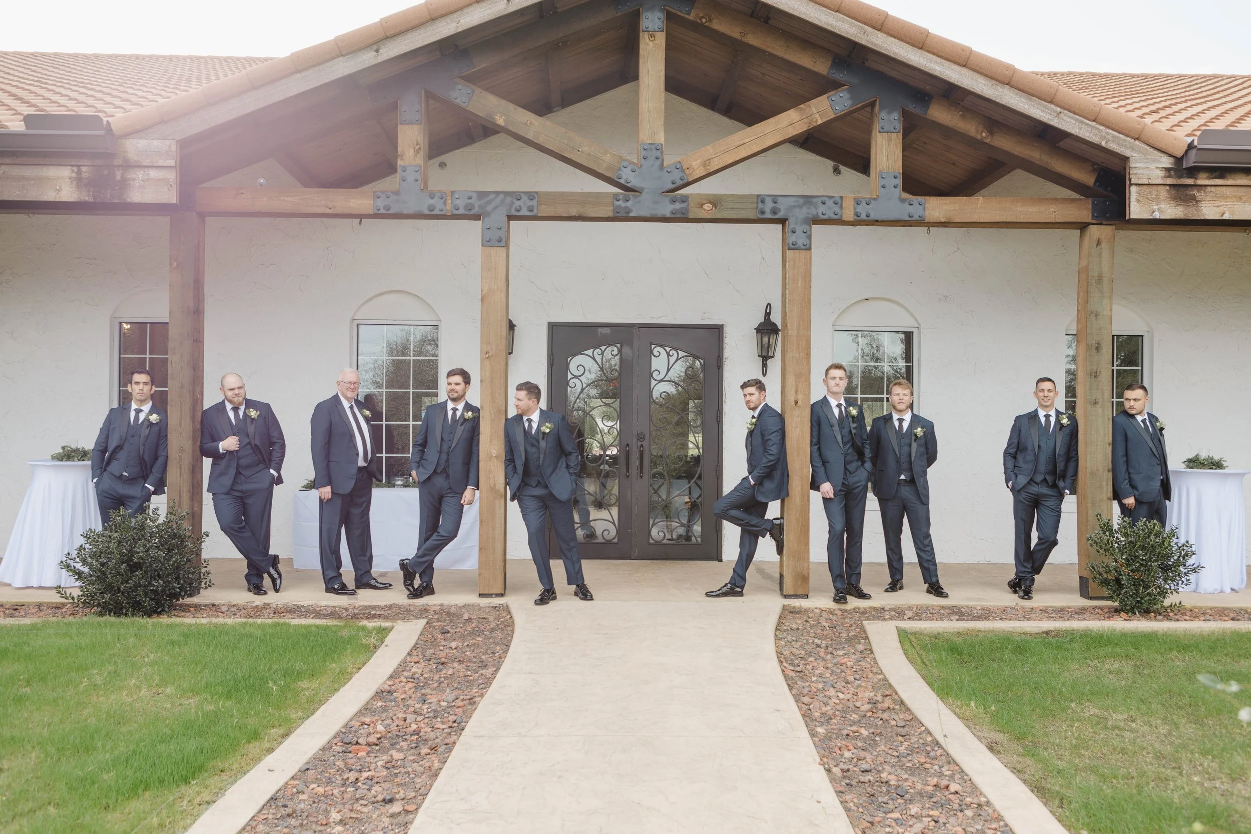 A group of nine men dressed in dark suits and ties standing outside in front of a house entrance with an arched black door with decorative wrought iron. The men are standing or leaning casually, some with hands in pockets or arms crossed, and they ar