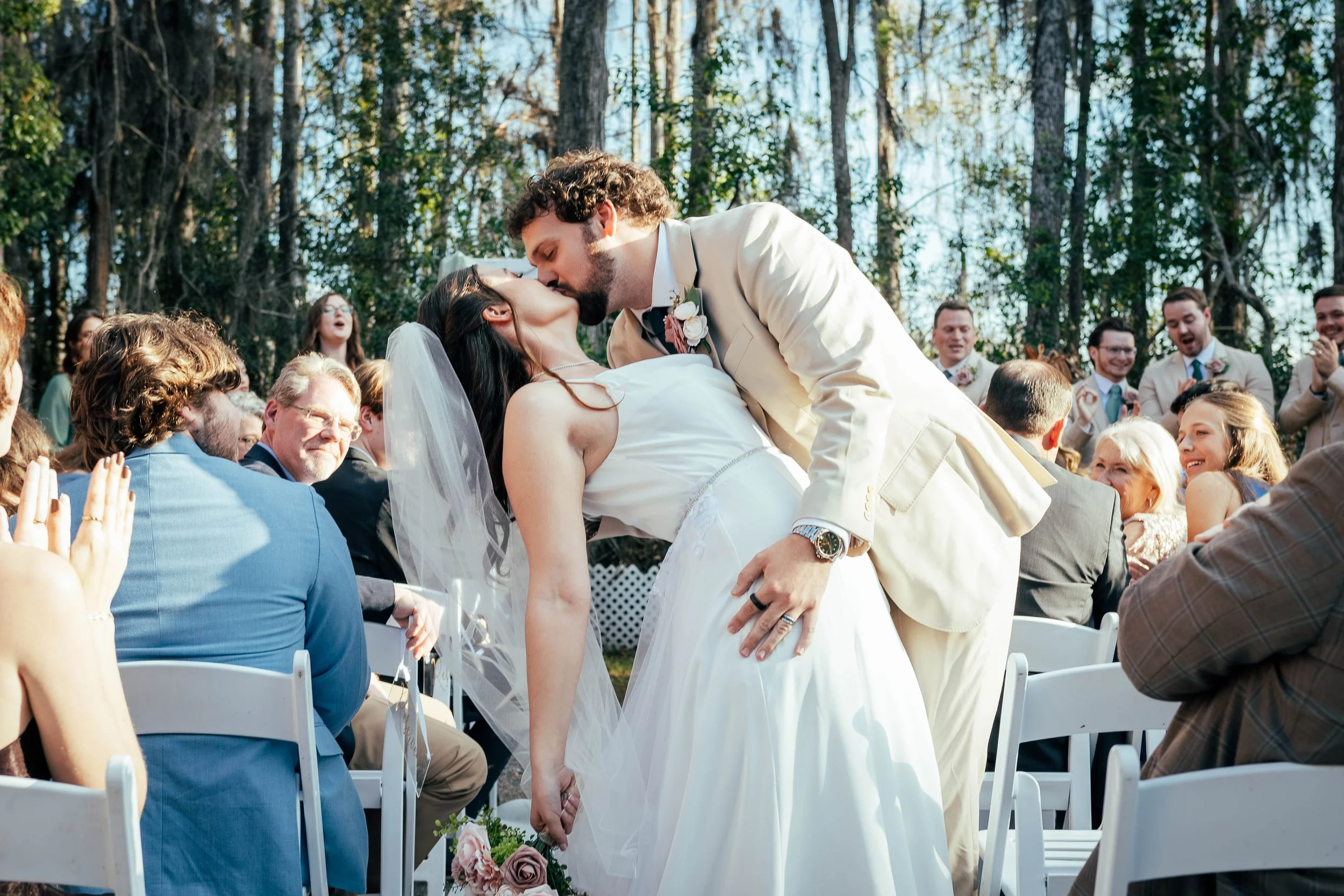 A newly married couple is sharing a kiss at an outdoor wedding ceremony surrounded by friends and family.