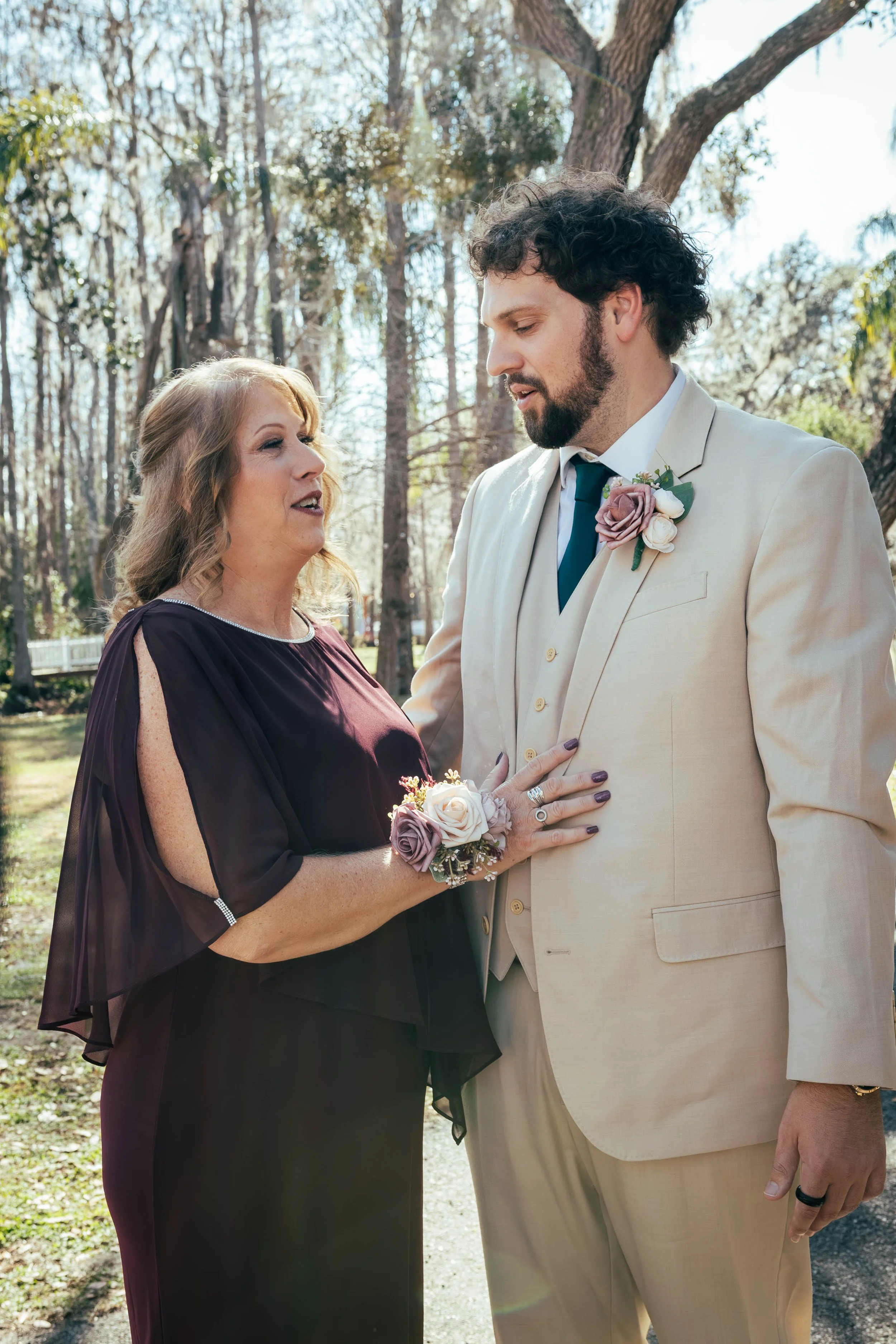 A woman and a man holding hands during a wedding ceremony outdoors in a wooded area. The woman is wearing a dark dress and a flower corsage, and the man is wearing a light-colored suit with a boutonniere.