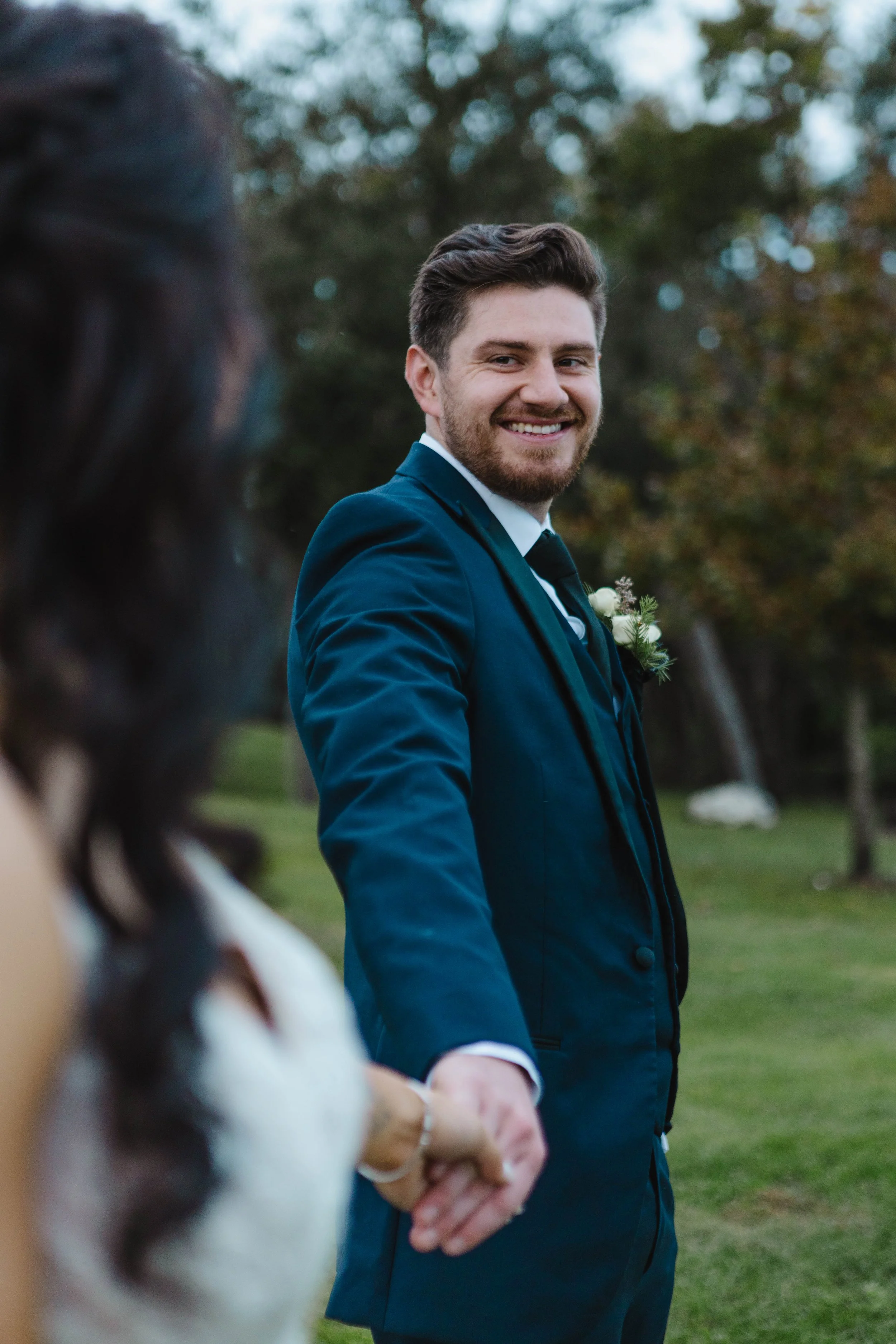 A smiling man in a blue suit and tie standing outdoors during daytime, holding hands with a woman, with blurred trees in the background.