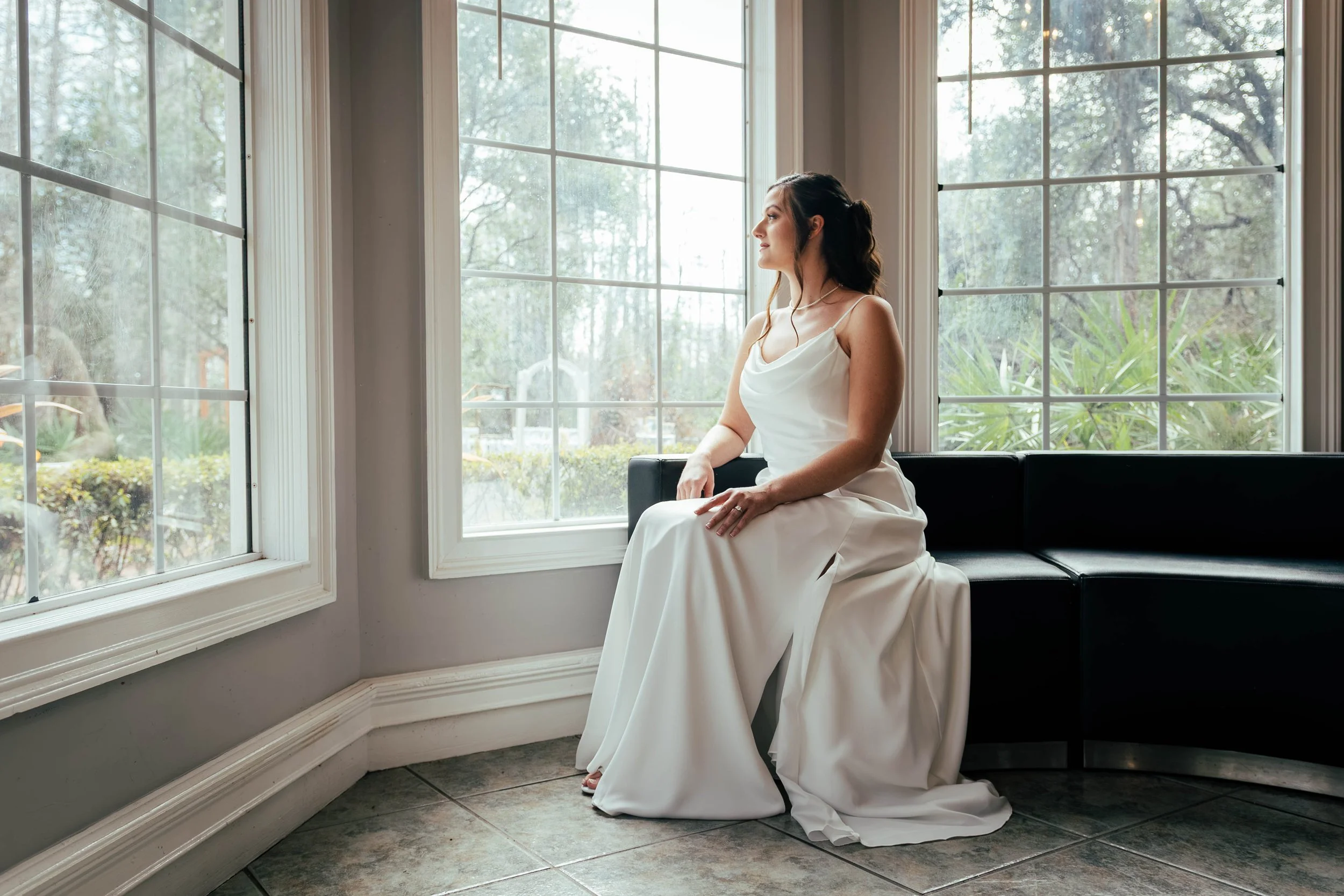 A woman in a white wedding dress sitting on a black bench by large windows with a garden view, softly lit by natural light.