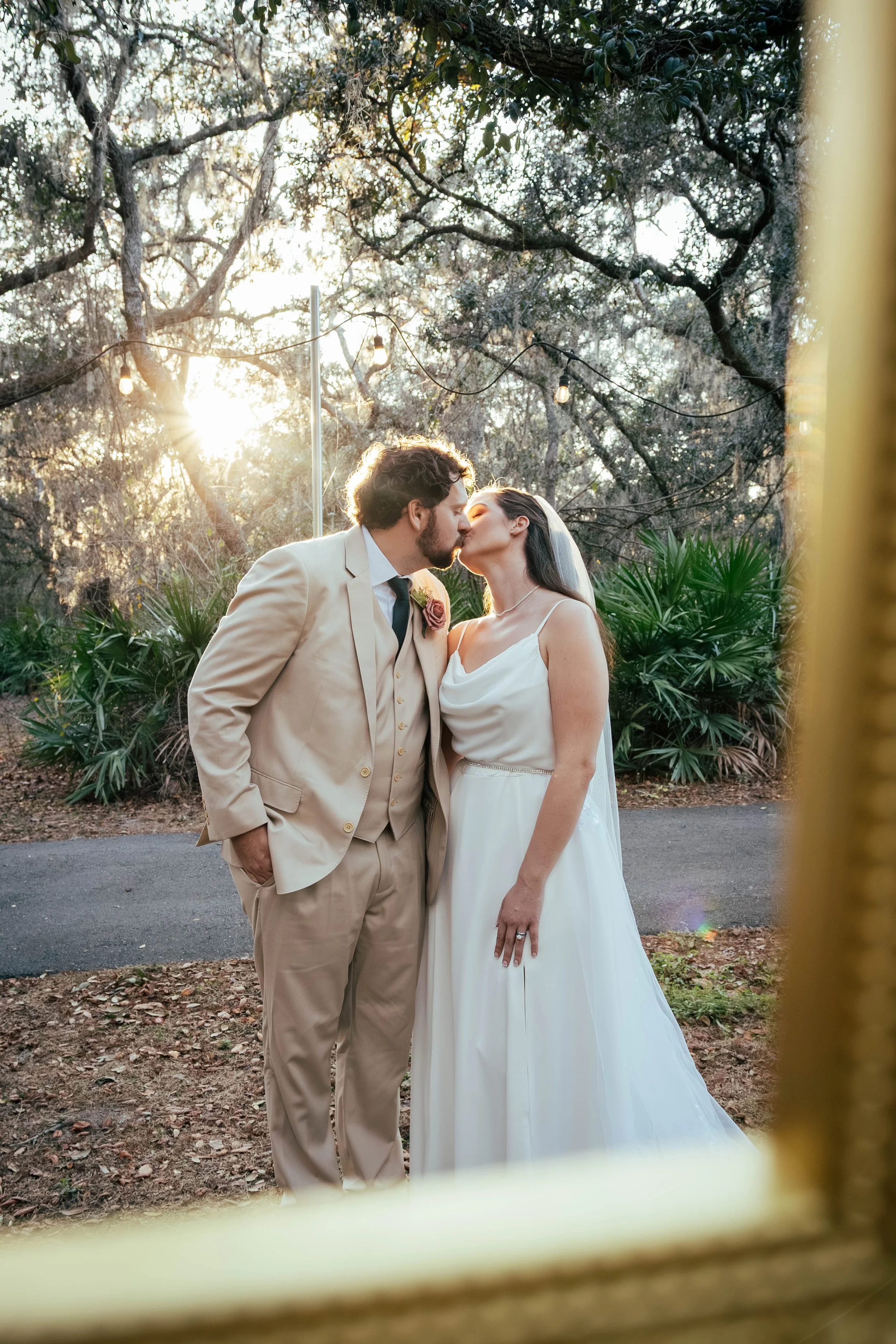 A bride and groom kissing outdoors during sunset, framed by a yellow fence in the foreground with trees and string lights in the background.
