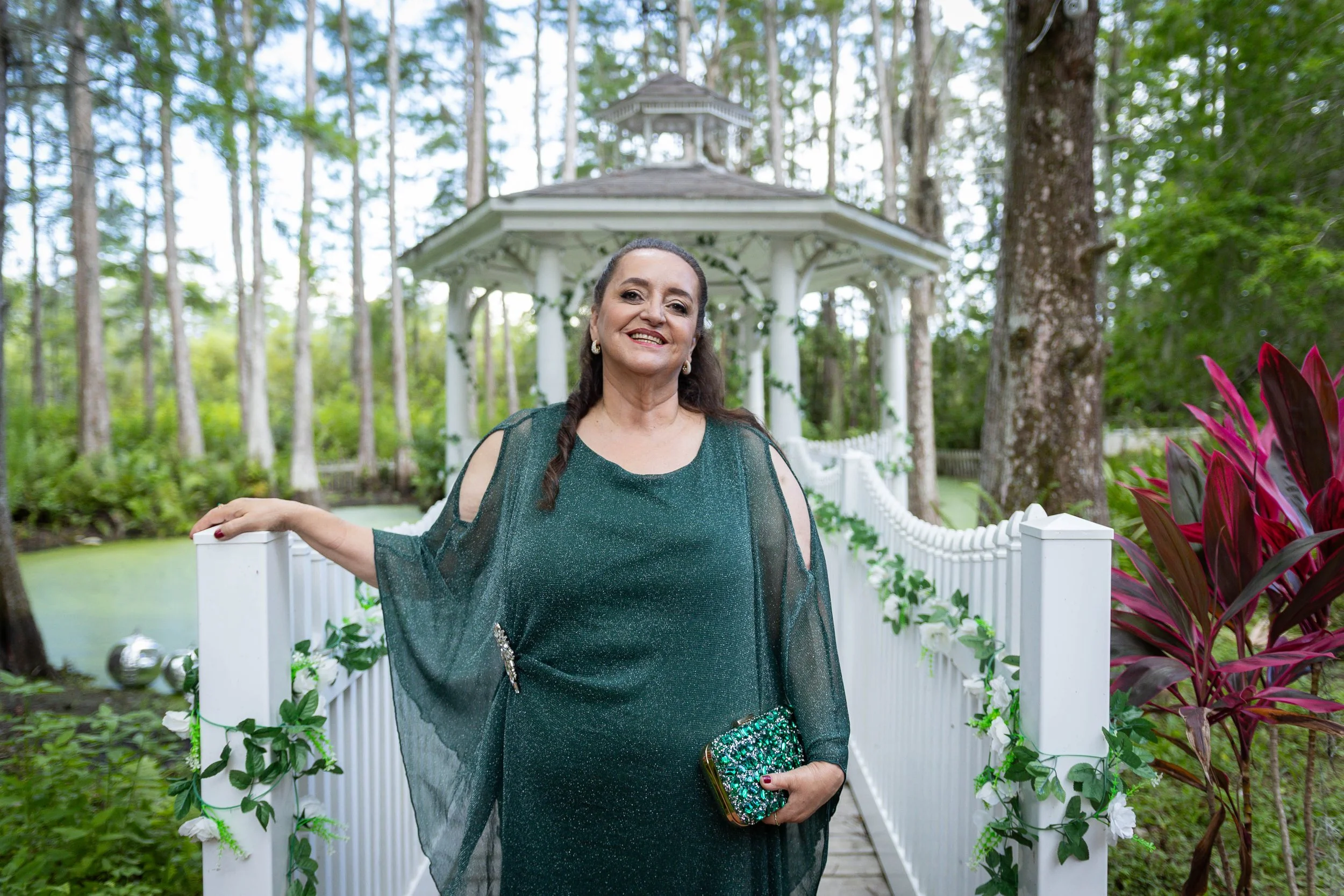 A smiling woman with dark hair in a braid, wearing a dark green, embellished dress and sheer overlay, holding a sparkly green clutch, standing on a white bridge in a lush garden with tall trees and vibrant plants.