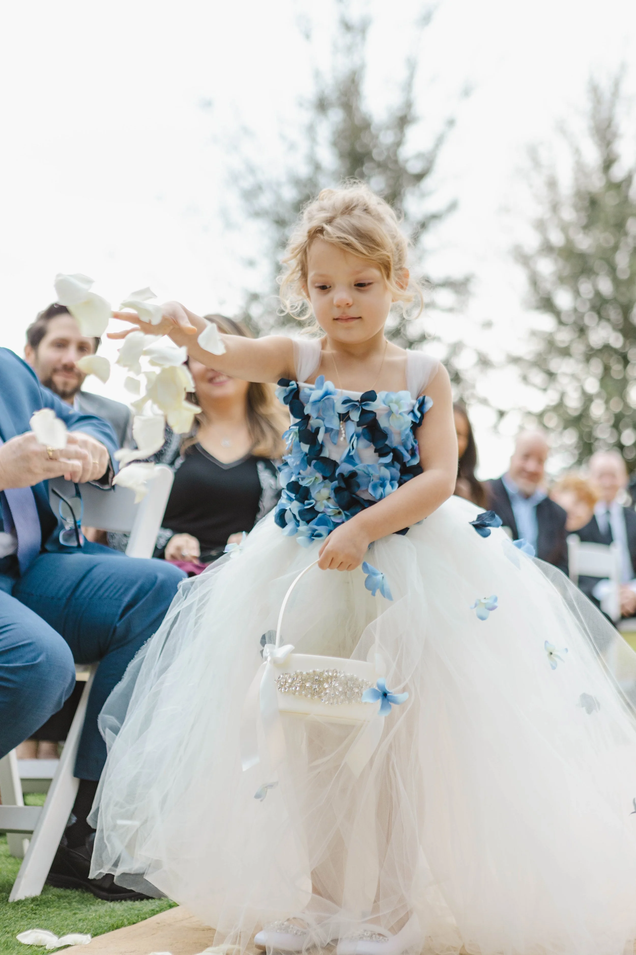 A young girl wearing a white and blue flower dress sitting outdoors during a wedding ceremony, with flower petals on her dress and a white basket on her lap, surrounded by adults in formal attire.