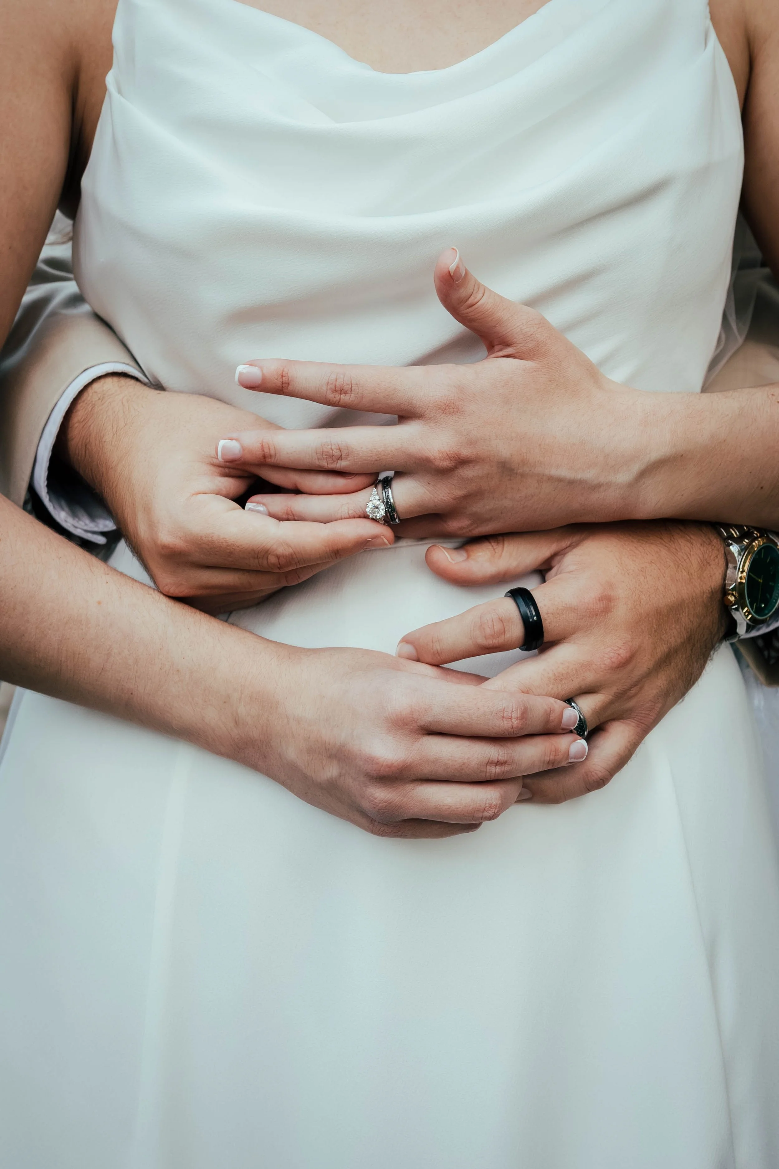 Close-up of a couple's crossed hands with wedding rings, embracing the woman in a white dress.