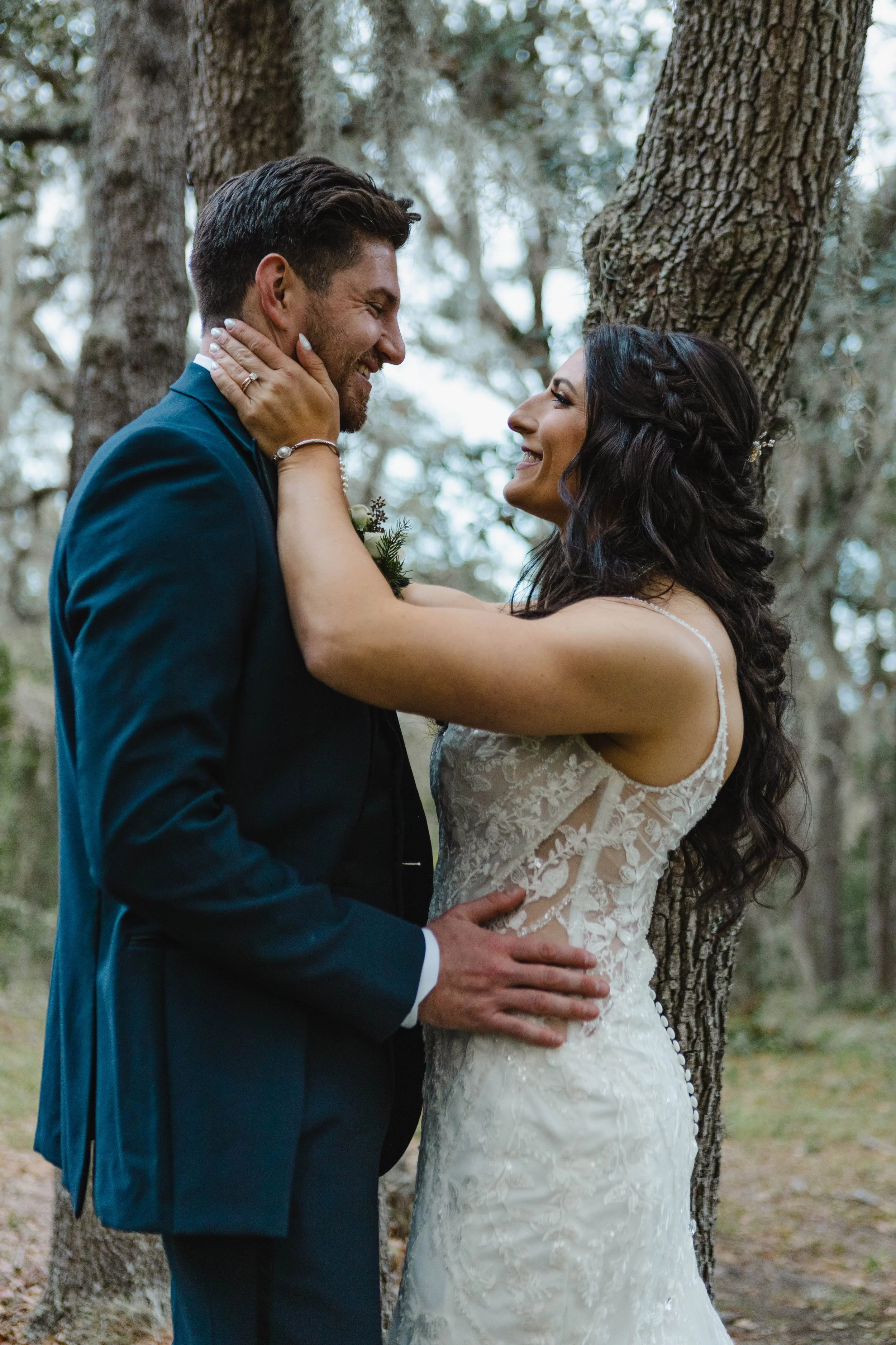 A newlywed couple in wedding attire stands close to each other outdoors, with a tree in the background. The woman touches the man’s face, and they both smile warmly.