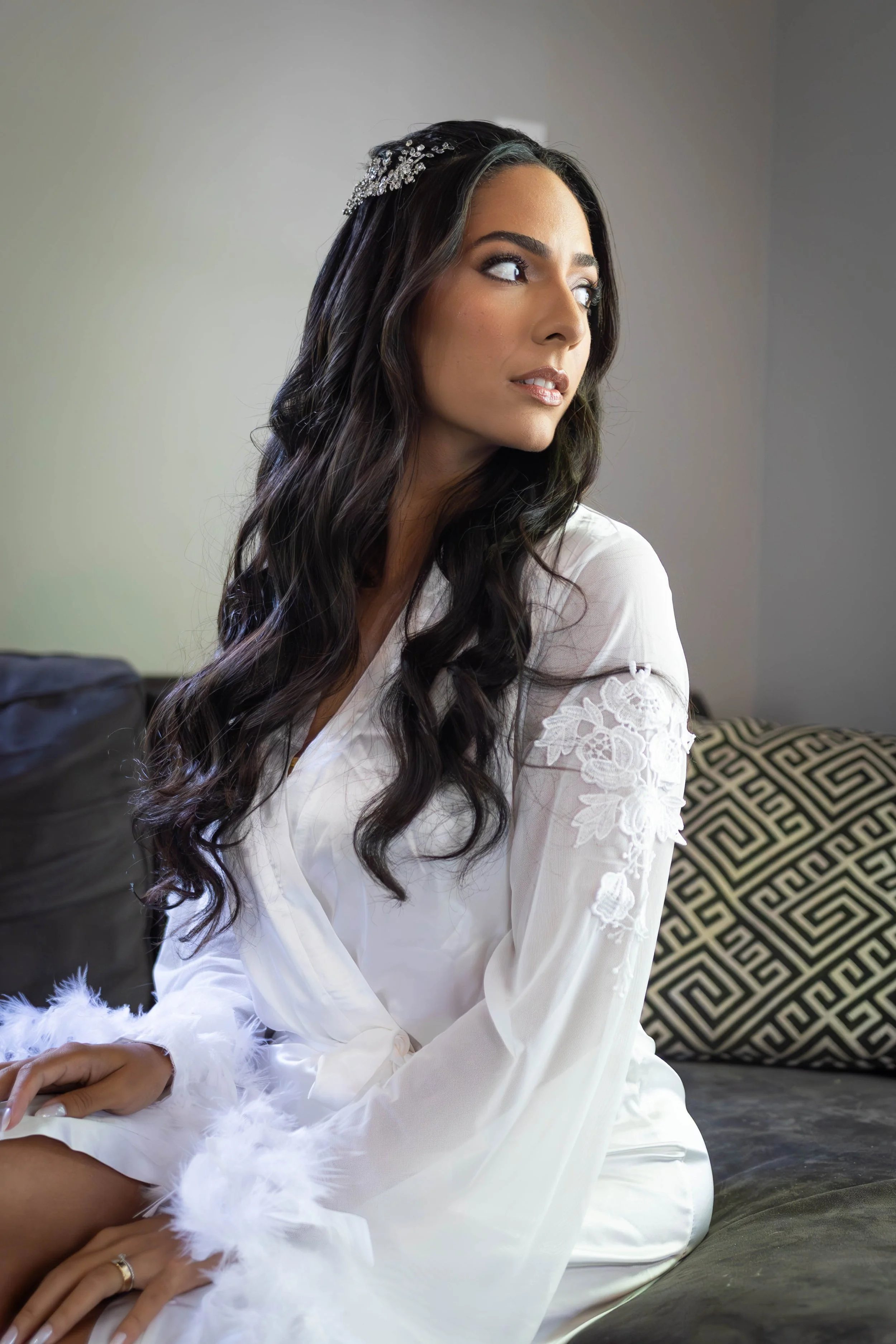 A woman with long dark wavy hair, wearing a white lace dress and a decorative hairpiece, sitting indoors on a couch with patterned pillows in the background.