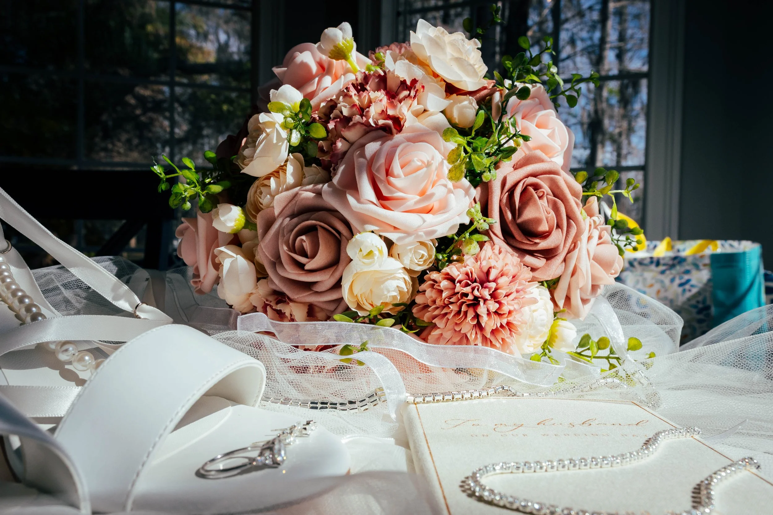 Close-up of a bridal bouquet with cream, peach, and burgundy roses along with greenery, placed on a table with wedding accessories including rings, jewelry, pearl necklace, and lace fabric, in natural light.