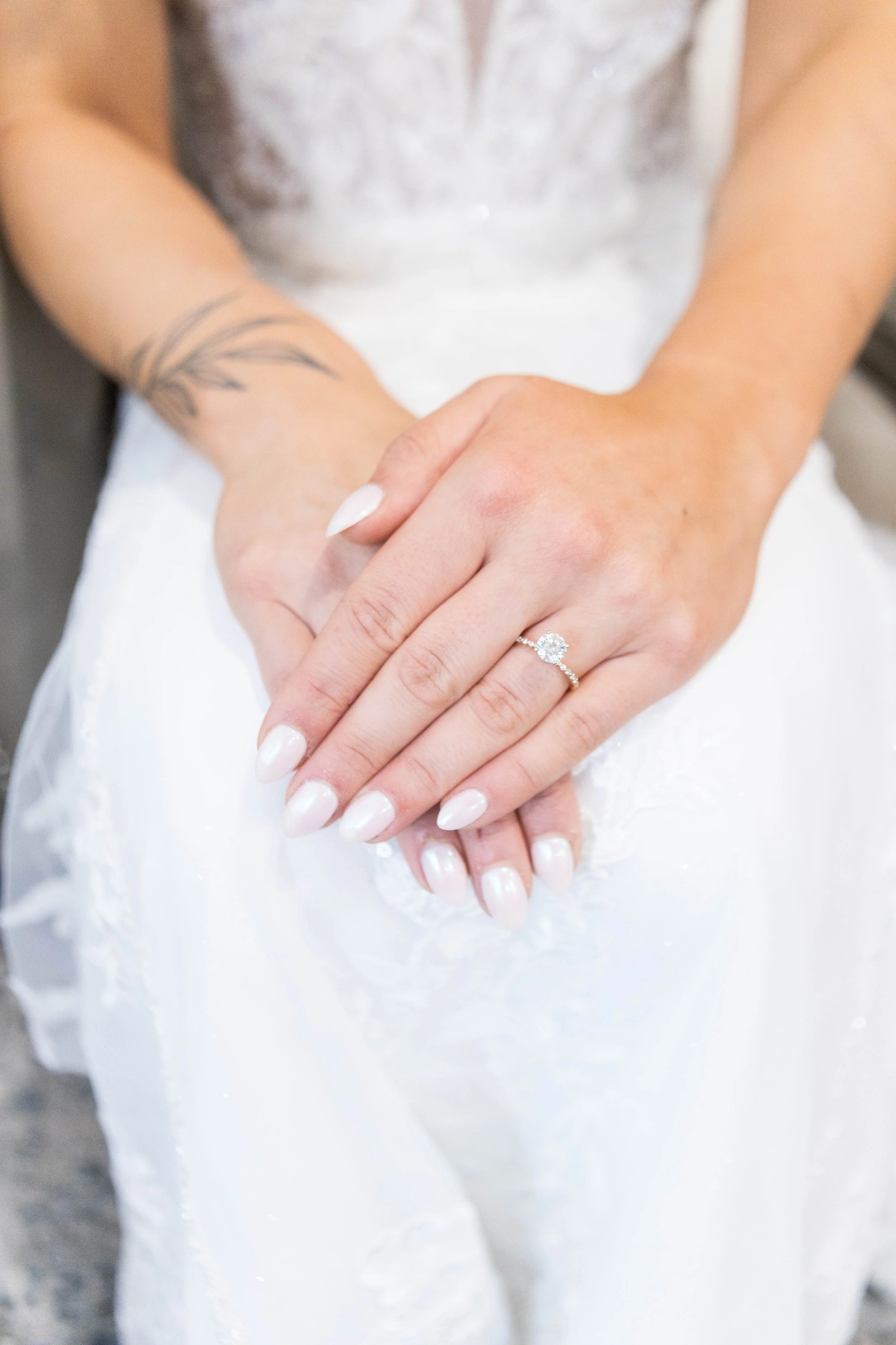 Close-up of a person's hands showing an engagement ring with a large round diamond on their left ring finger, with their other hand resting on top of their hands. The person has a black tattoo on their left wrist and is wearing a light-colored dress 