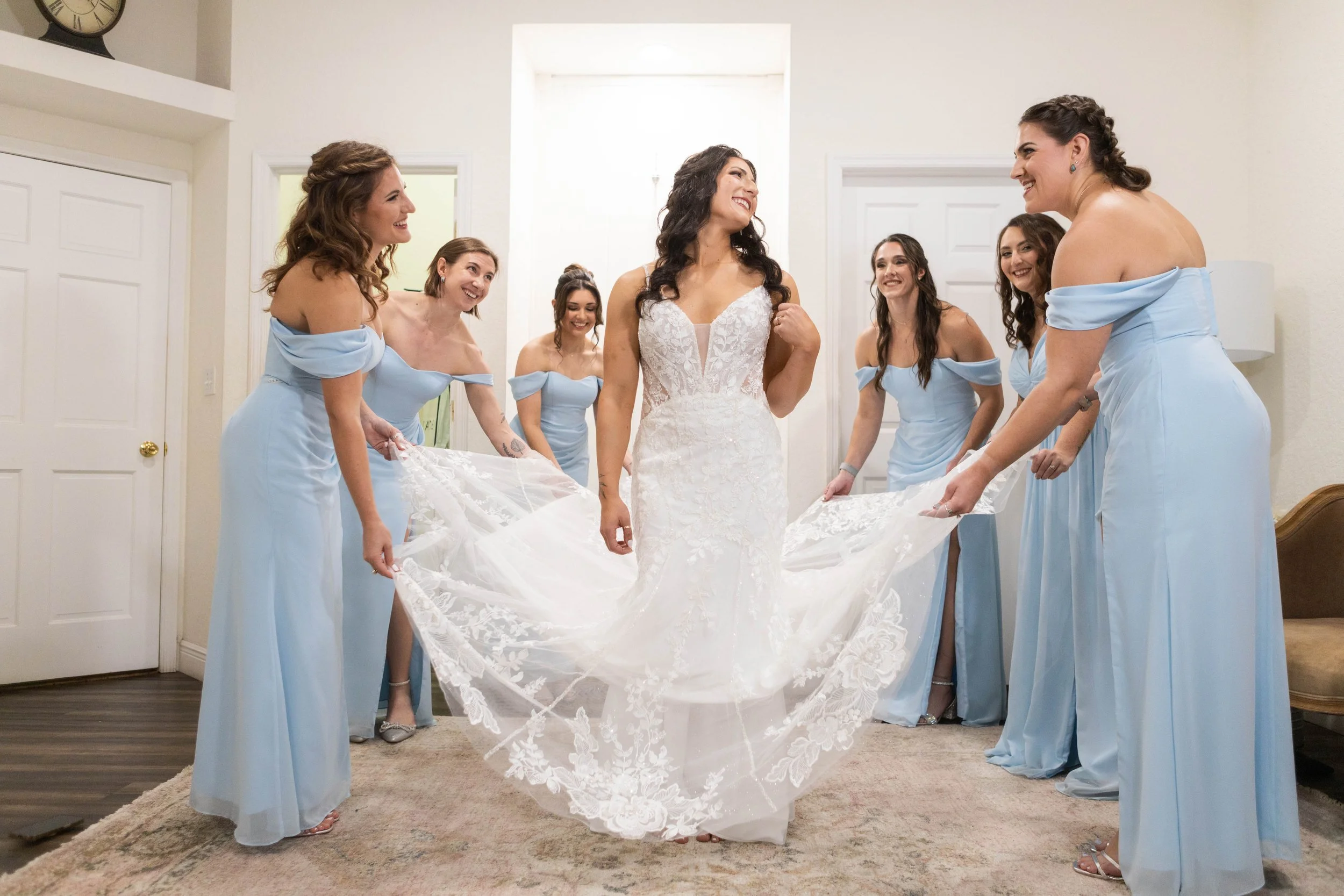 Bride in a white lace wedding gown surrounded by six bridesmaids in light blue dresses, smiling and holding the train of the gown.