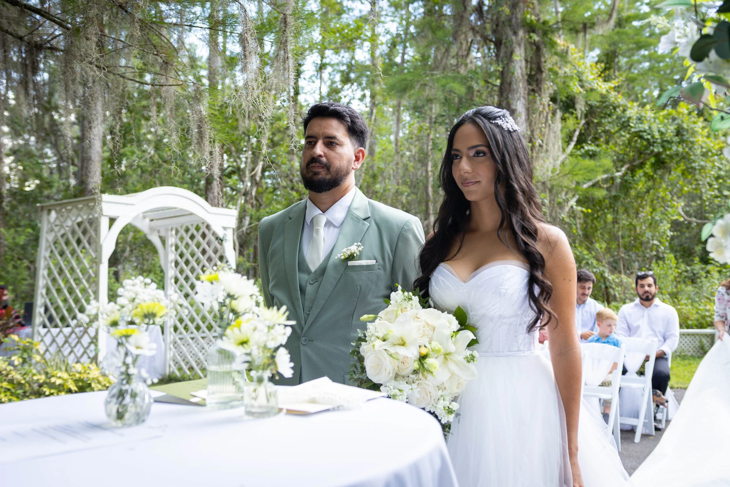 A bride and groom standing outdoors at a wedding ceremony in a lush green wooded setting. The bride wears a white strapless wedding gown and holds a bouquet of white flowers, while the groom wears a light gray suit with a white shirt and tie. Guests 