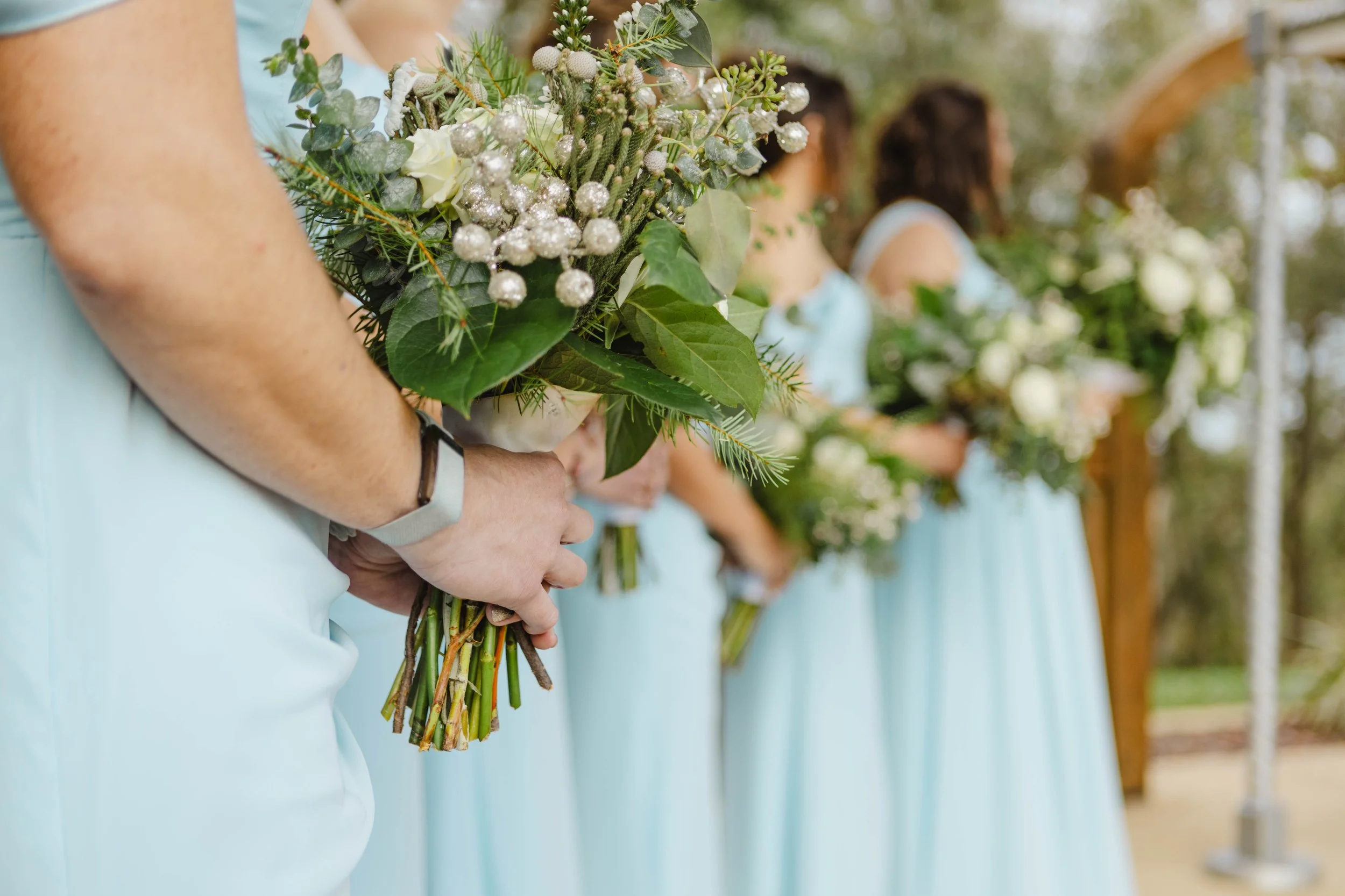 A row of women dressed in light blue outfits holding bouquets of flowers and greenery, standing outdoors near a pool or lake with trees and wooden structures in the background.
