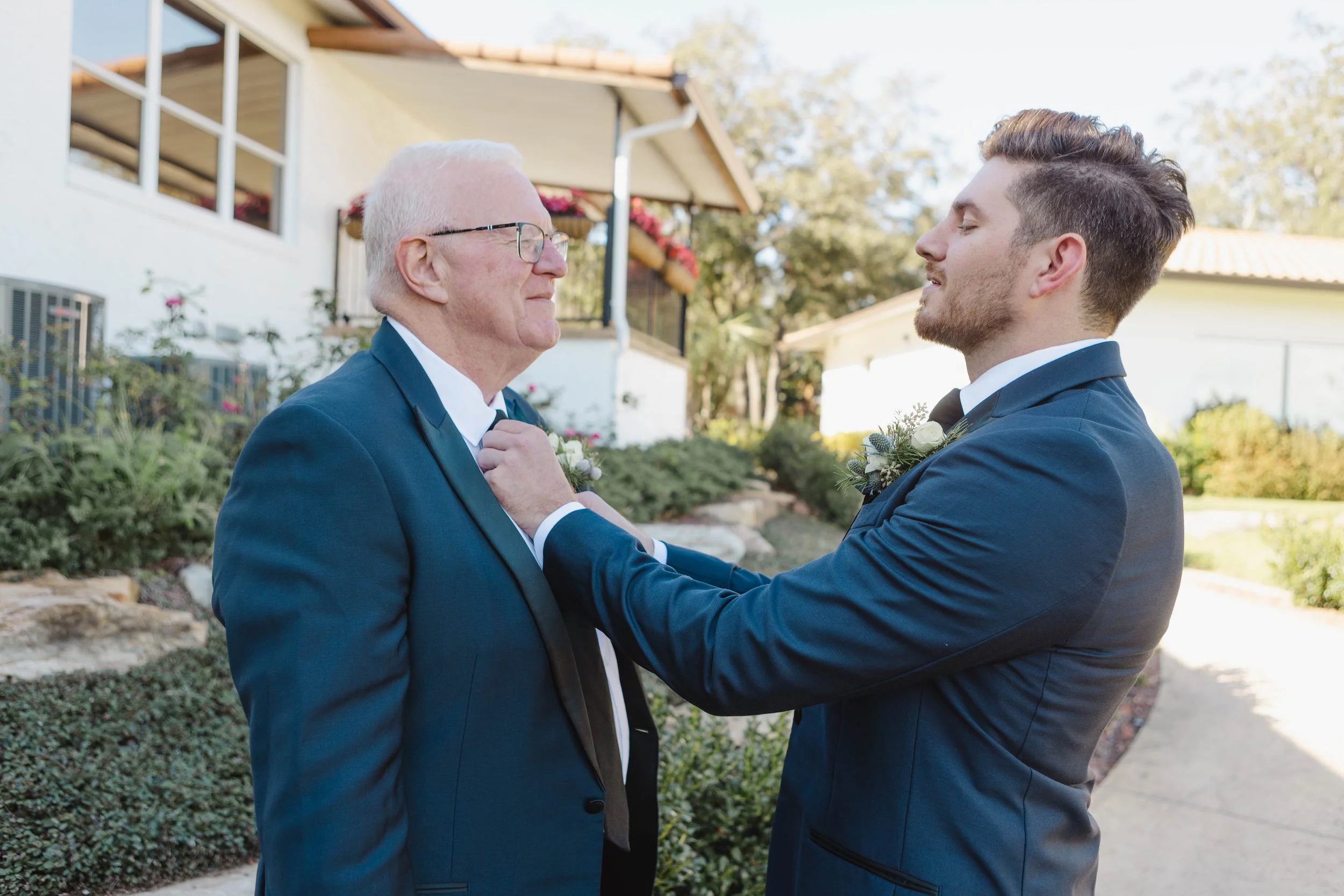 An elderly man and a young man in formal suits facing each other outdoors, with the elderly man adjusting the young man's bow tie, in front of a house with trees and plants.