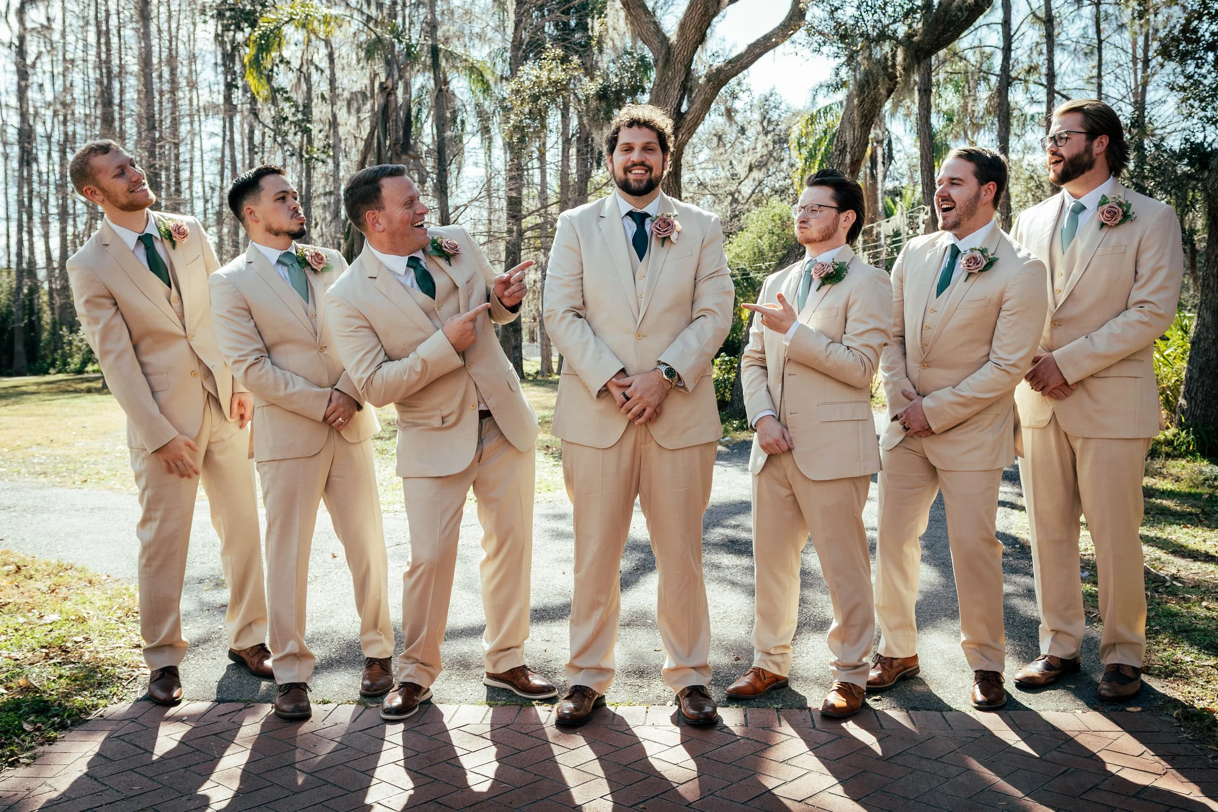 A group of men dressed in beige suits with blue and pink boutonnieres, standing outdoors in a wooded area, smiling and talking to each other.