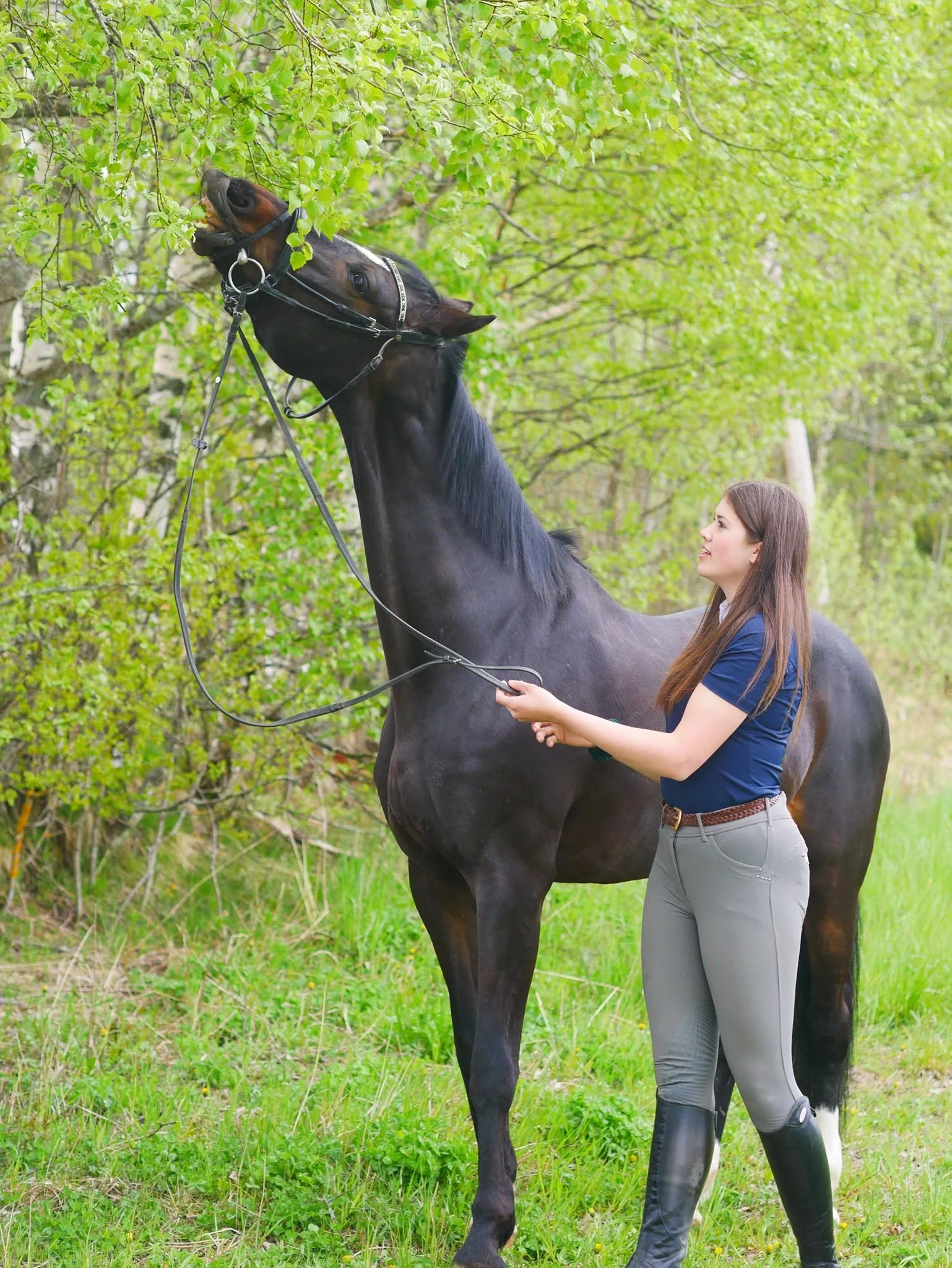 En kvinne og en svart hest med brunt i hodet, står i en grønn skog.