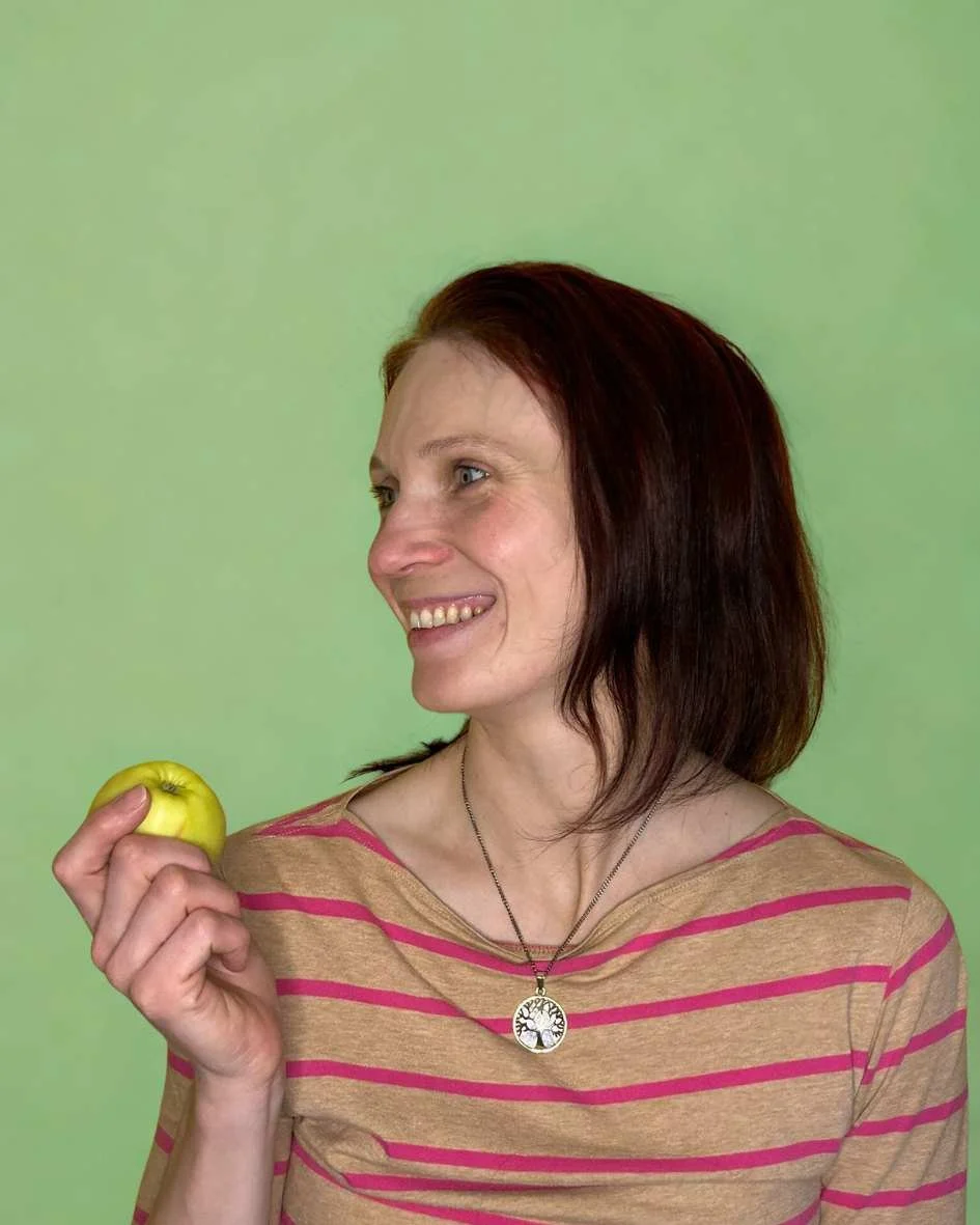 Nutritional therapist smiling and holding an apple, inviting to join natural health community