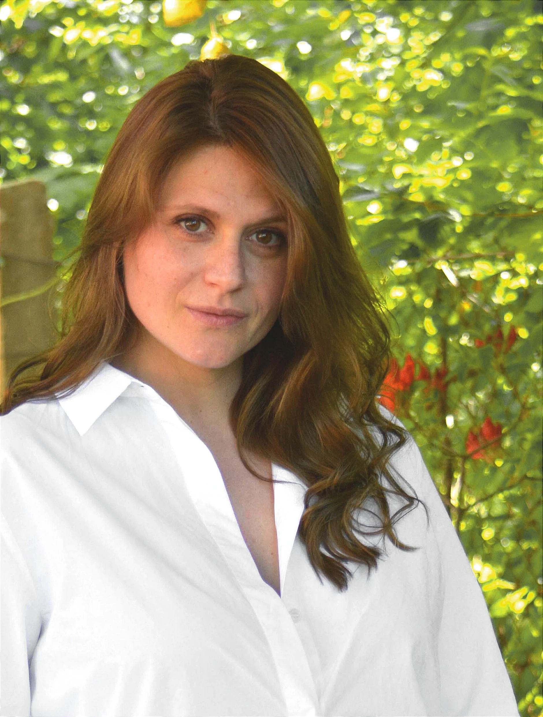 Porträt einer Frau mit braunen, gewellten Haaren, die weiße Bluse trägt, vor einem grünen, bewaldeten Hintergrund bei Tageslicht.