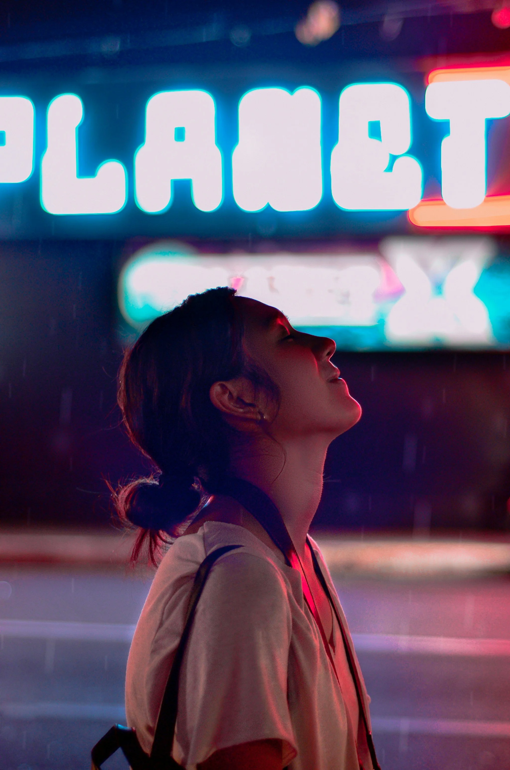 Eine junge Frau steht bei Nacht mit geschlossenen Augen vor einer Neonreklame. Das Licht der Neonwerbung spiegelt sich auf ihrem Gesicht.