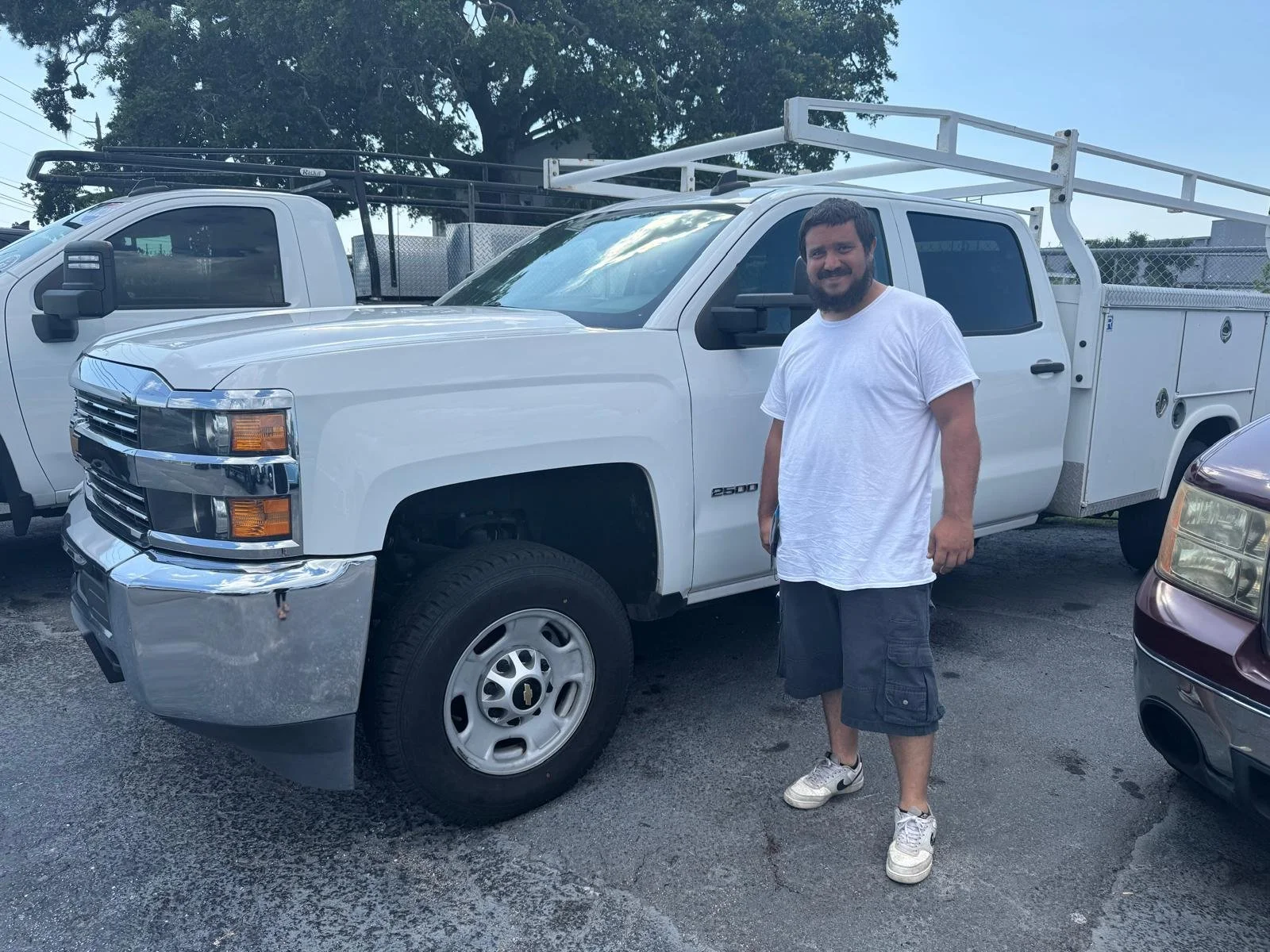 A man with a beard and wearing a white t-shirt, cargo shorts, and sneakers standing next to a white utility pickup truck with a ladder rack on top, parked at Sunshine Auto in Pinellas Park FL 33781