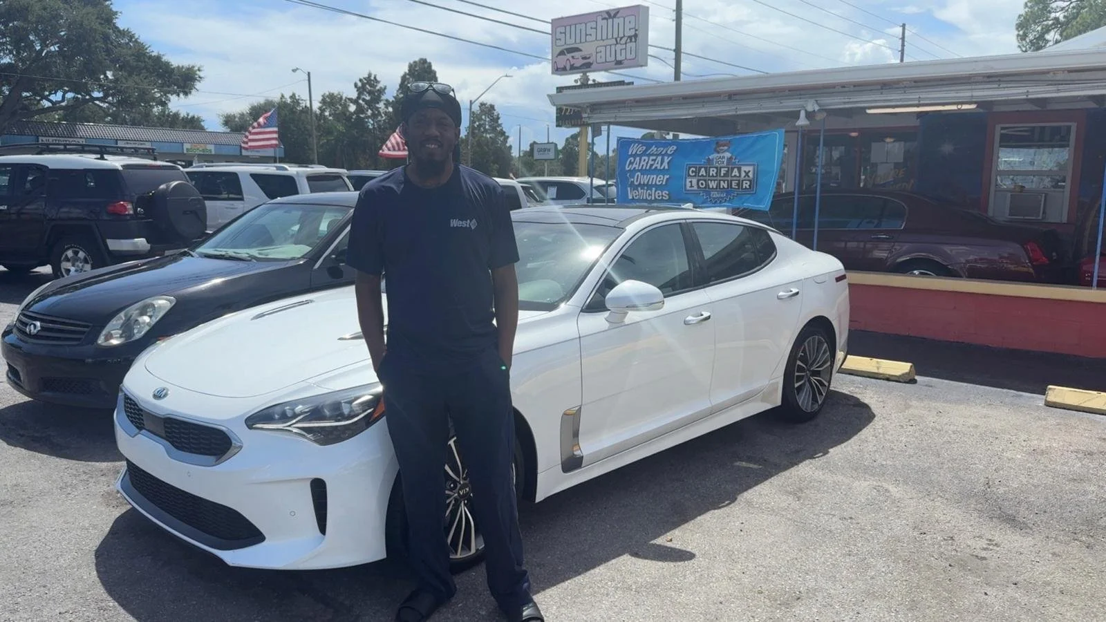 A man standing next to a white car in a car dealership lot. There are several other cars parked nearby, with a sign that reads 'Sunshine Auto' and banners advertising CARFAX vehicle history reports.