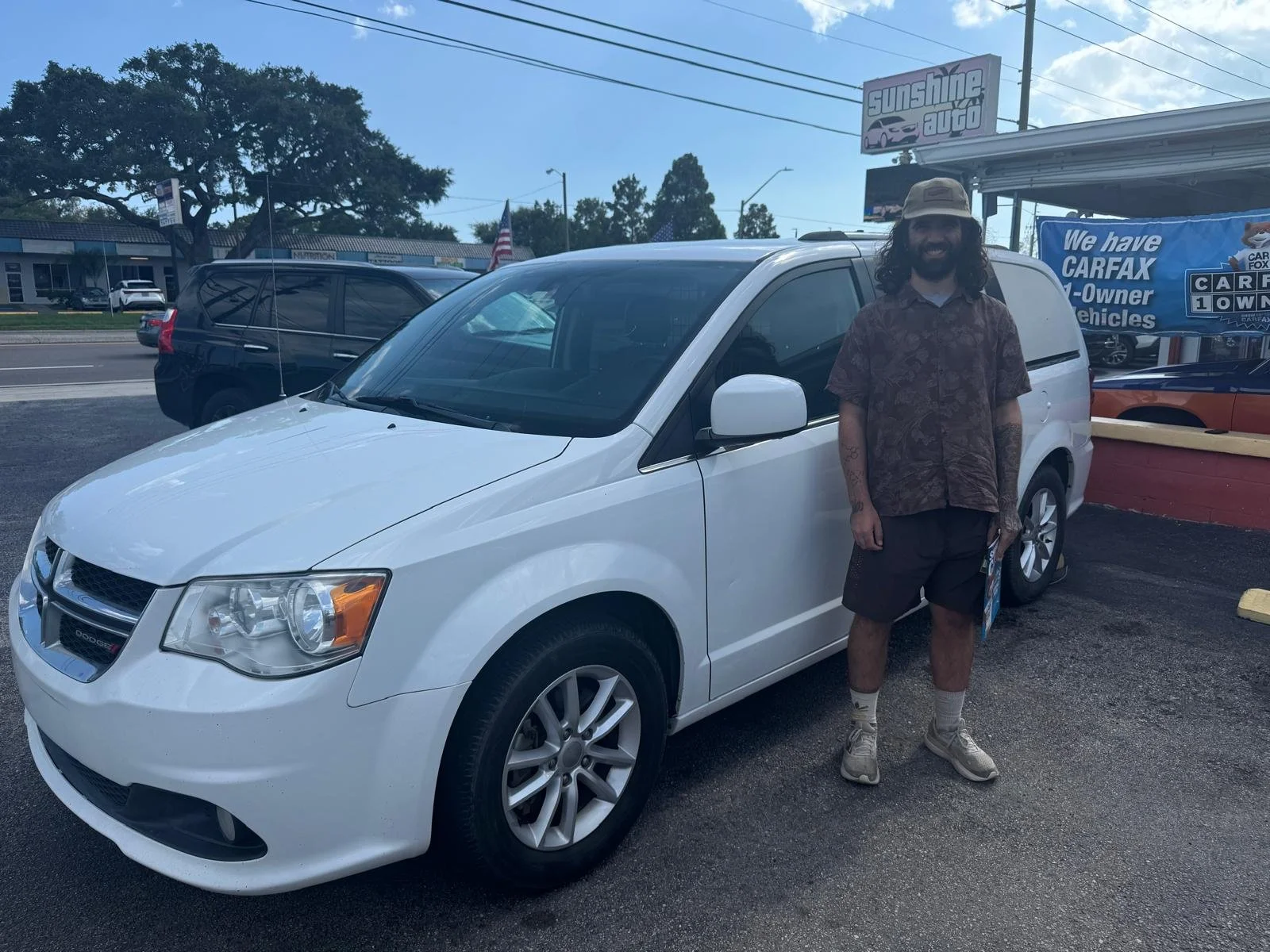 A man with long curly hair, wearing a hat, a patterned shirt, and shorts, standing next to a white minivan at Sunshine Auto in Pinellas Park FL 33781
