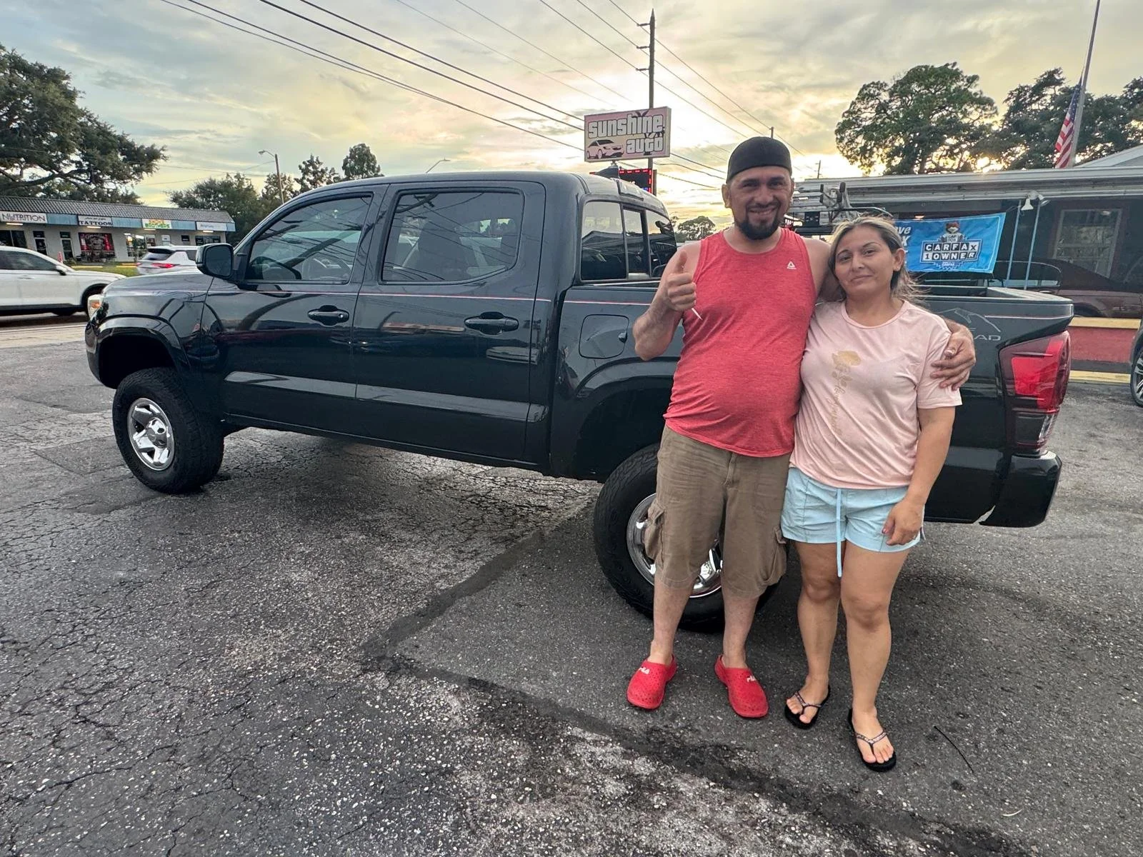 A man and woman standing arm in arm in front of a black pickup truck, smiling at the camera during sunset, in a parking lot with a car dealership in the background.