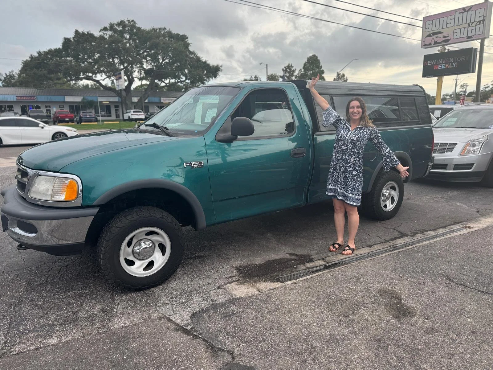 A woman in a black and white dress standing next to a teal Ford F-150 pickup truck at a car dealership lot, smiling with her arm raised.